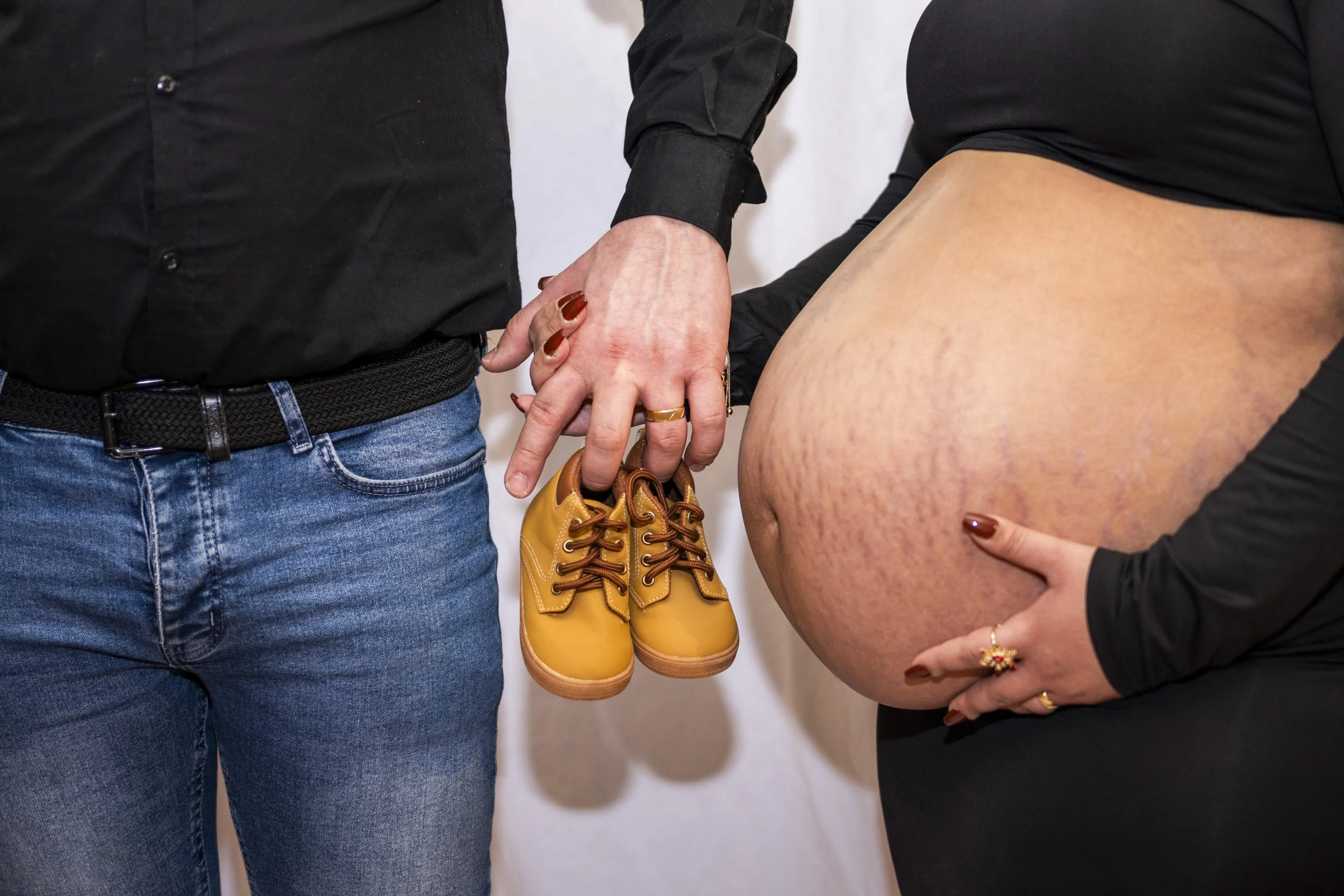 A pregnant woman in black shirt holding her belly and touching her thigh, standing next to a person in blue jeans and a black shirt whose hand is holding tiny yellow baby shoes with brown laces.