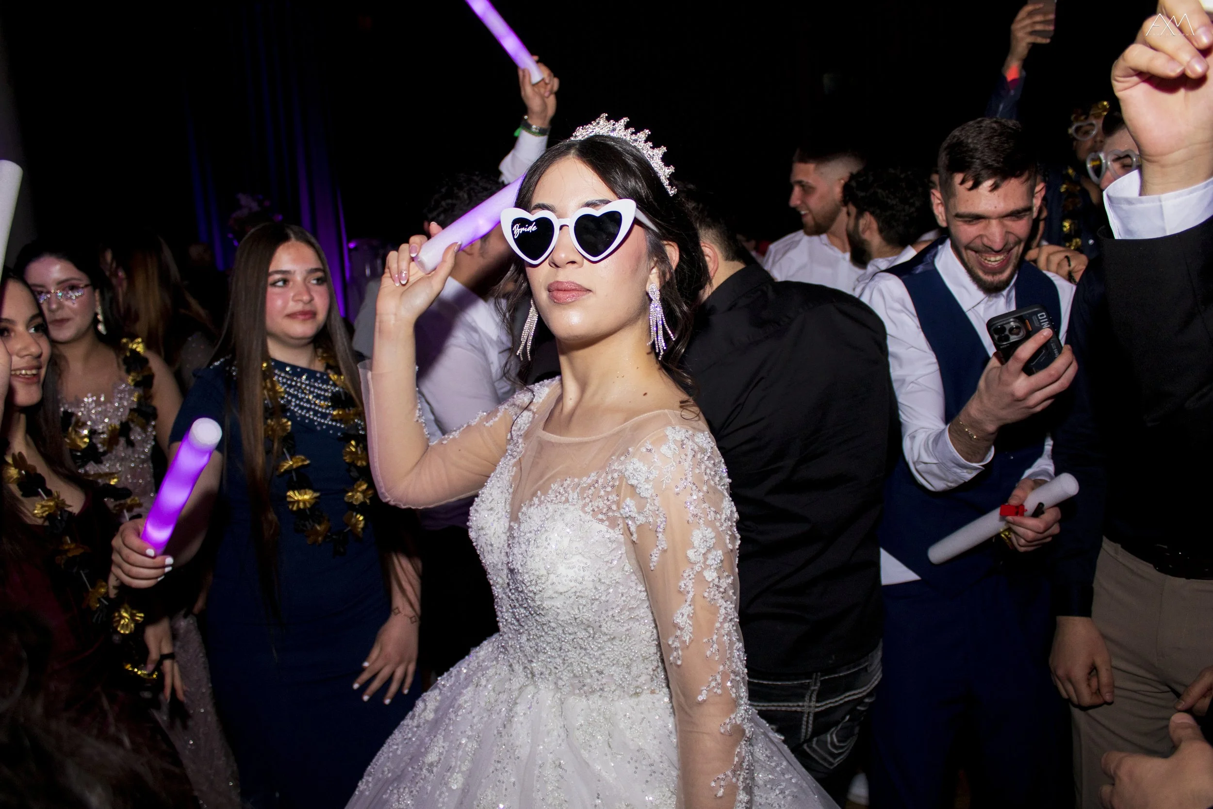 A bride in a white lace wedding gown and tiara, wearing heart-shaped sunglasses with the word "bride" on them, poses at a celebration with smiling guests around her. The guests are holding glow sticks and phones, and are dressed in formal and festive