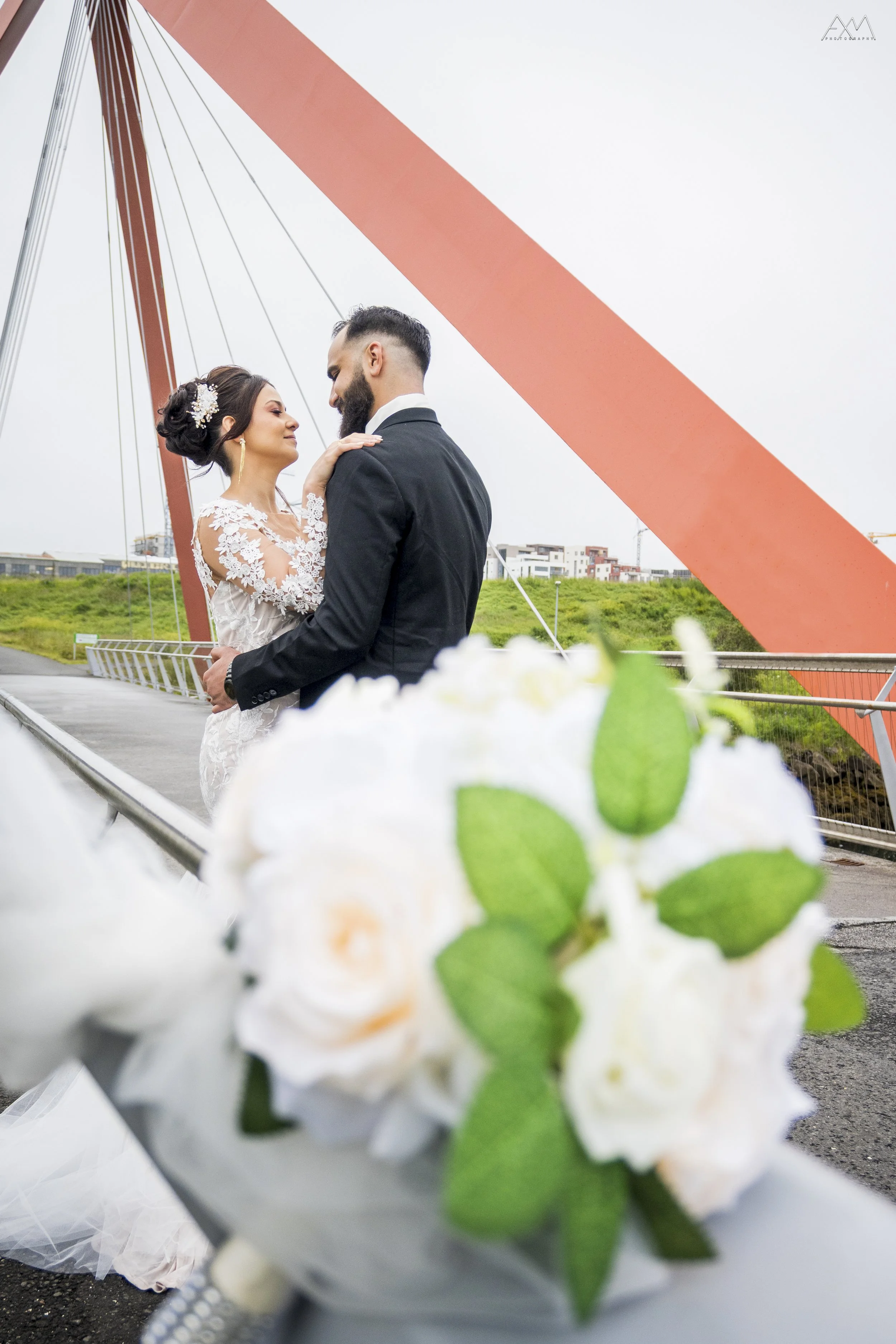 A bride and groom sharing an intimate moment on a bridge during their wedding. The bride wears a lace wedding dress and has an elaborate updo with floral accessories, while the groom is in a dark suit with a white shirt. The background shows a cloudy