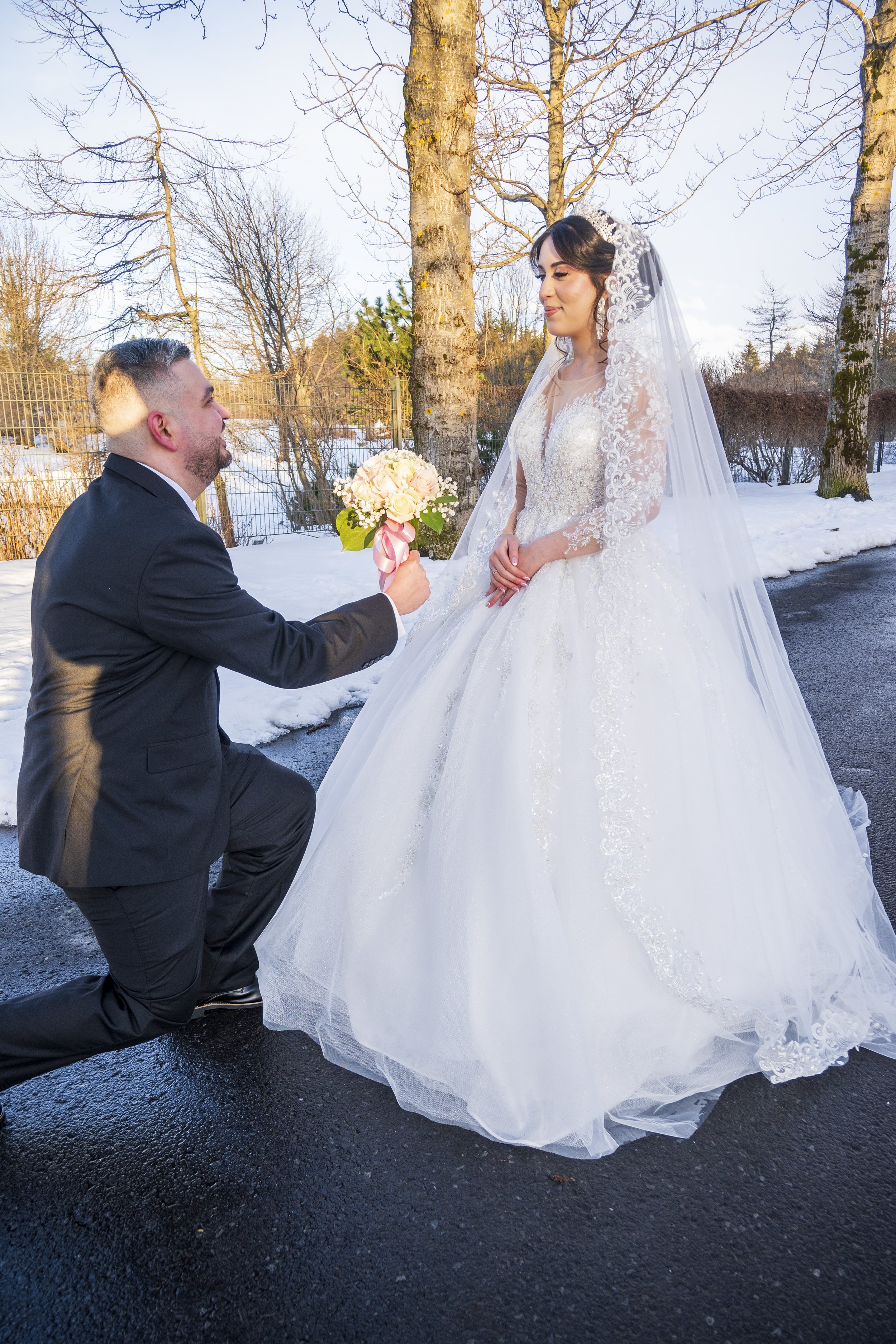 A man in a black suit is kneeling on one knee, holding a bouquet of white and pink flowers with greenery, and proposing to a woman in a white wedding gown with lace sleeves and a long train. The woman is looking tenderly at the man, outdoors with sno
