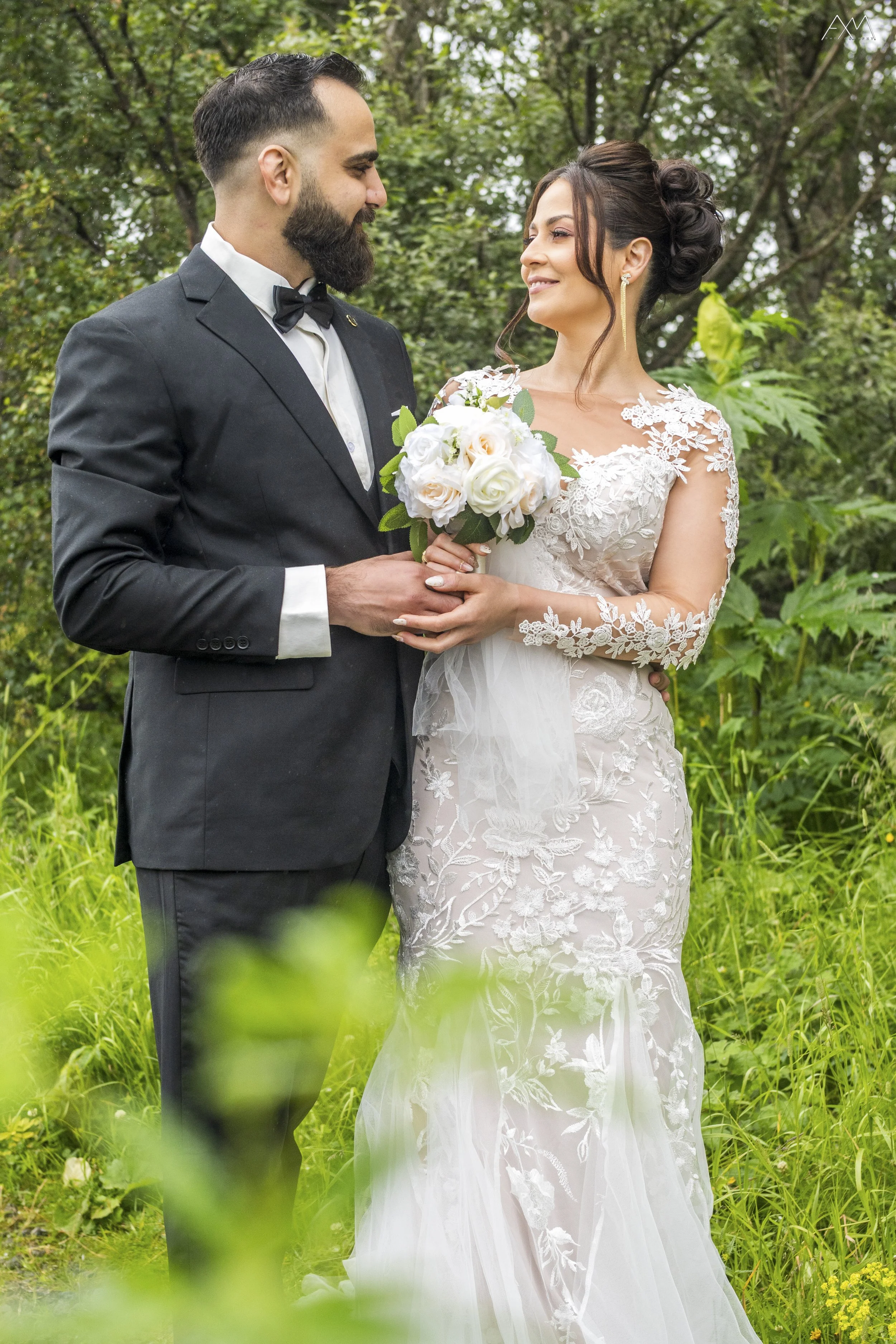 A bride and groom standing outdoors amidst greenery, looking at each other lovingly. The bride is holding a bouquet of white roses and wearing a lace wedding dress, and the groom is dressed in a black tuxedo with a bow tie.