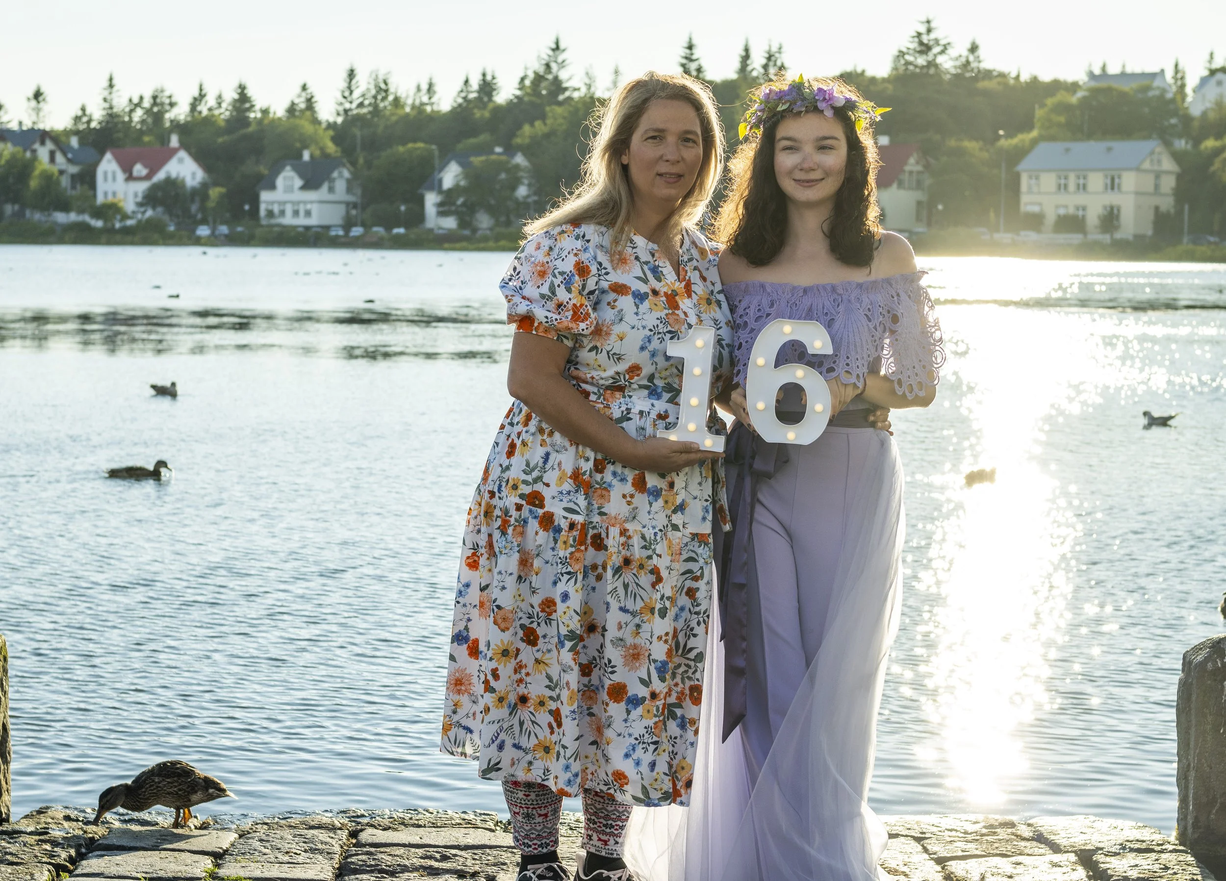 A mother and daughter standing by a lake at sunset, holding illuminated numbers '16' for a celebration, with ducks swimming nearby and houses in the background.