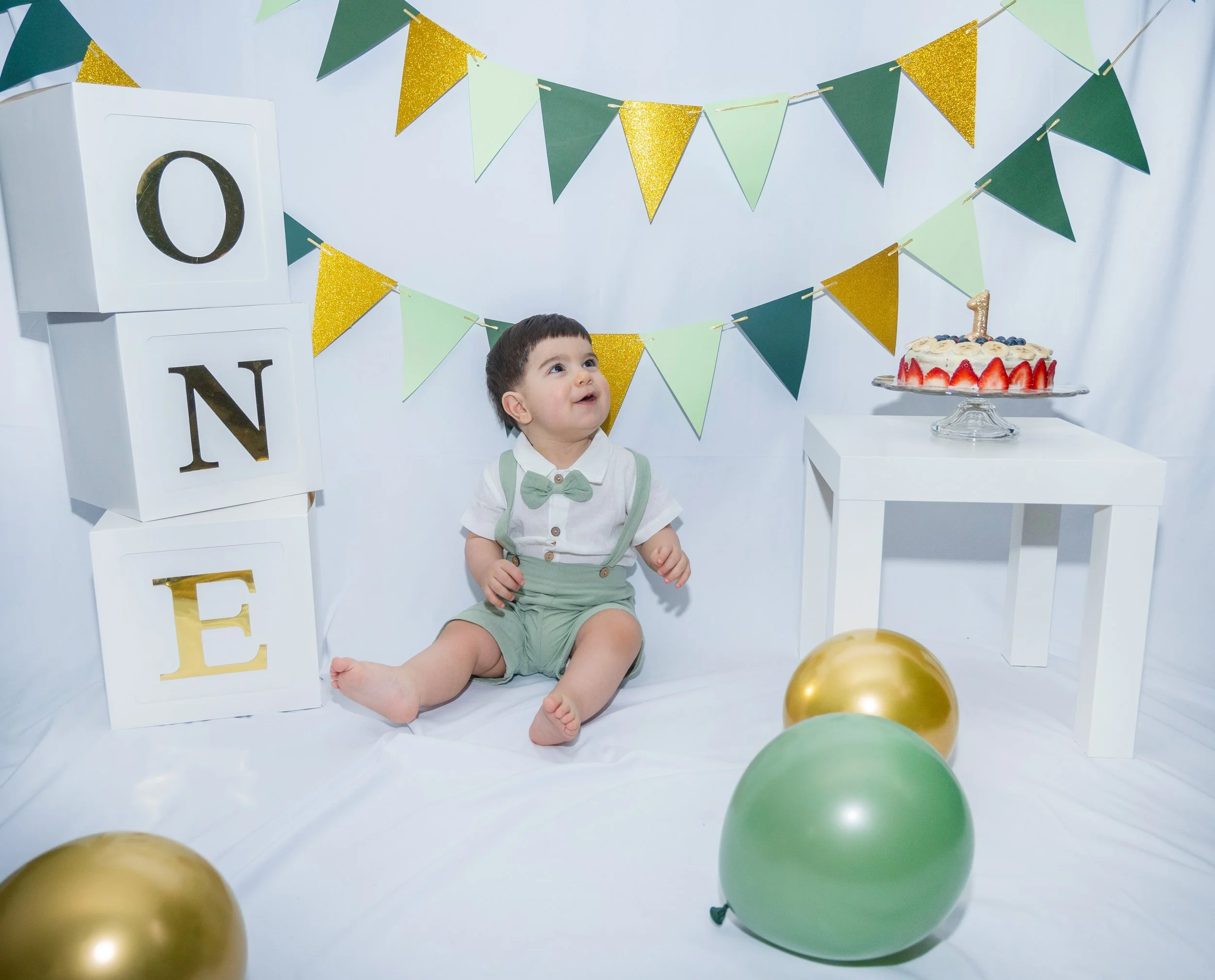 A young boy sitting on a white backdrop at a birthday celebration with a decorated cake, balloons, and green, gold, and light green pennant banners.