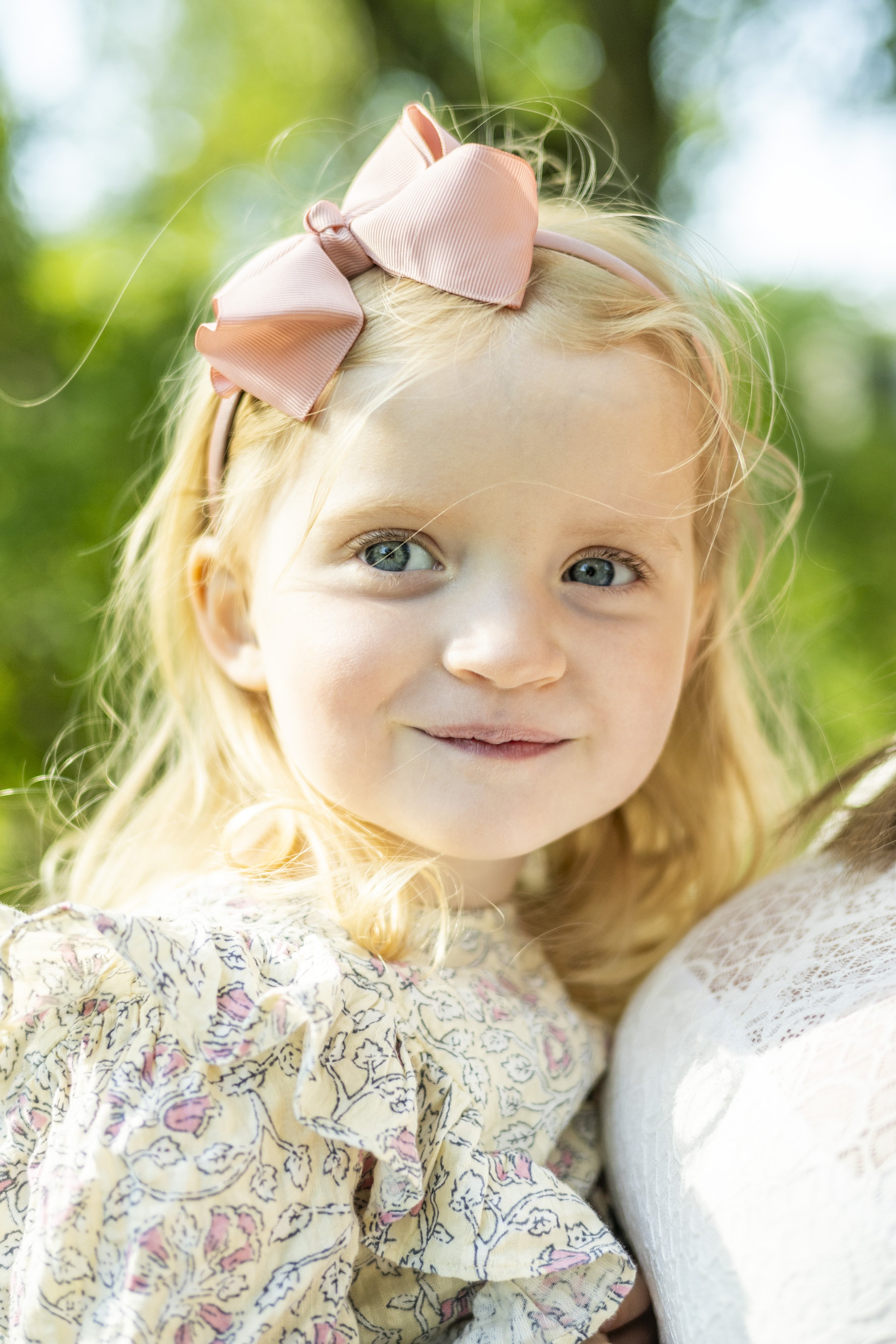A young girl with blonde hair, blue eyes, wearing a pink bow headband and a floral dress, smiling outdoors with blurred greenery in the background.