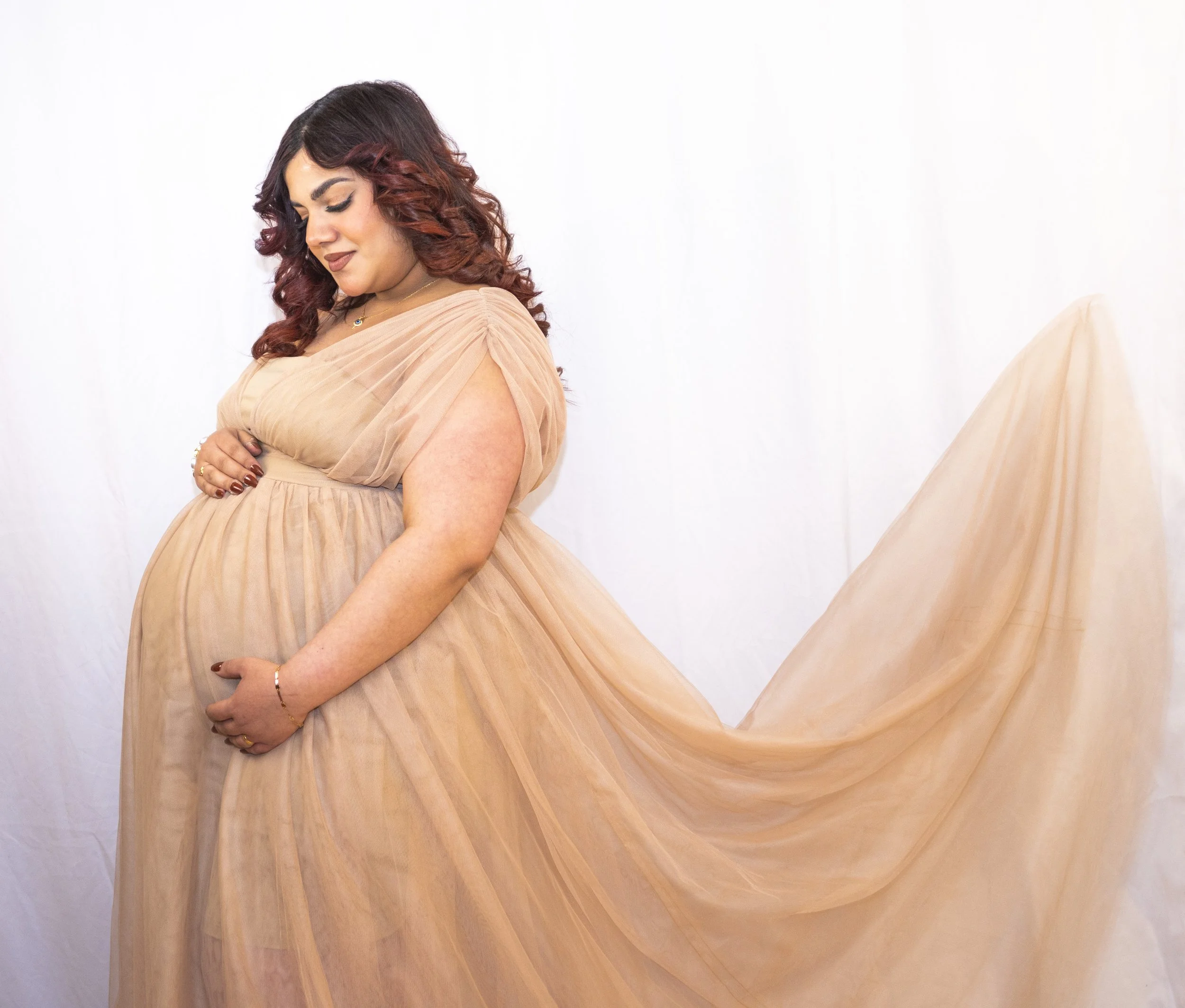 A woman with curly red-brown hair wearing a beige, pleated, off-shoulder dress, standing against a plain white background, with her eyes closed and hands crossed over her abdomen.
