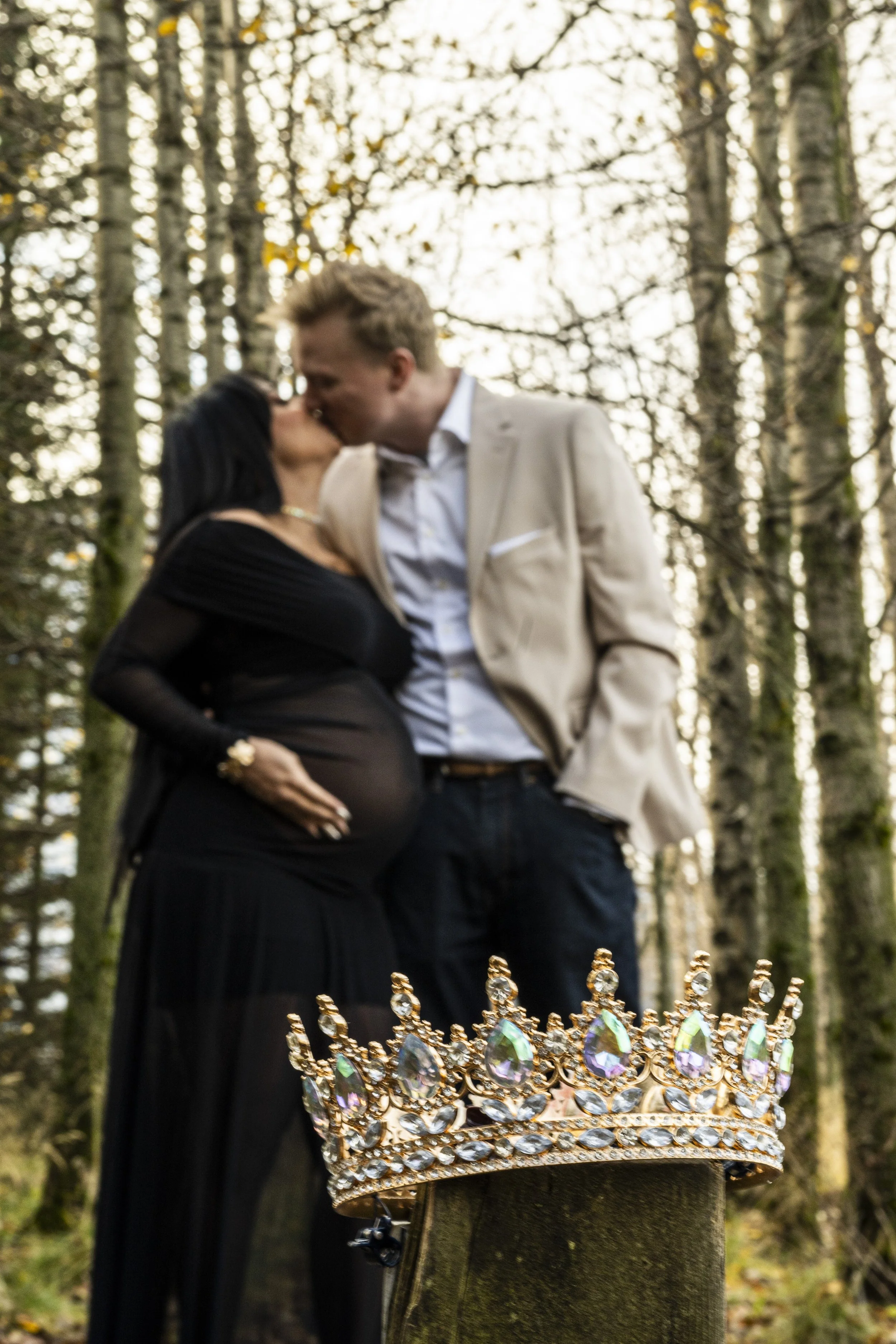 A couple kissing outdoors in a forest, with a gold and jewel-encrusted crown in the foreground.