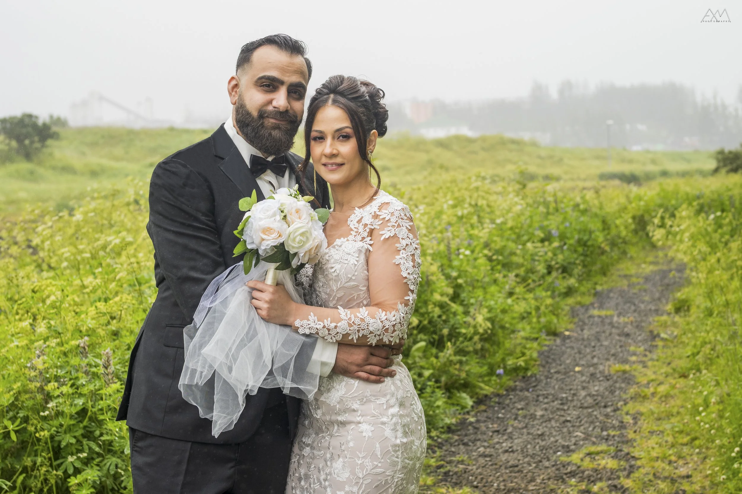 A bride and groom standing outdoors on a cloudy day, posing for a wedding photo. The groom is wearing a black tuxedo with a bow tie, and the bride is in a white lace wedding dress holding a bouquet of white roses and greenery. They are smiling and lo
