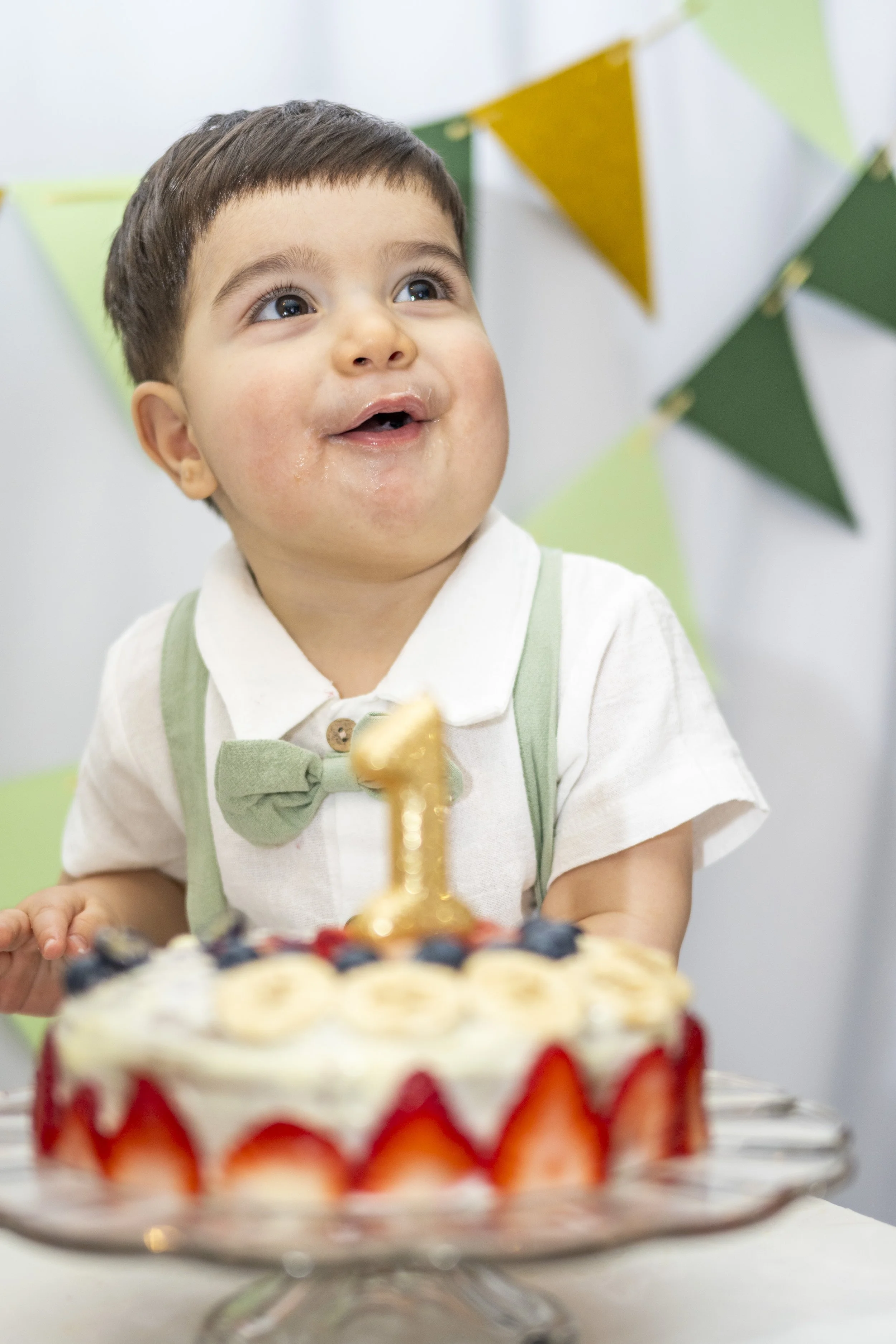 A young boy with short brown hair and big eyes, wearing a white shirt with a green bow tie and suspenders, sits in front of a birthday cake. The cake is decorated with strawberries and blueberries, and has a gold '1' candle on top. The background fea