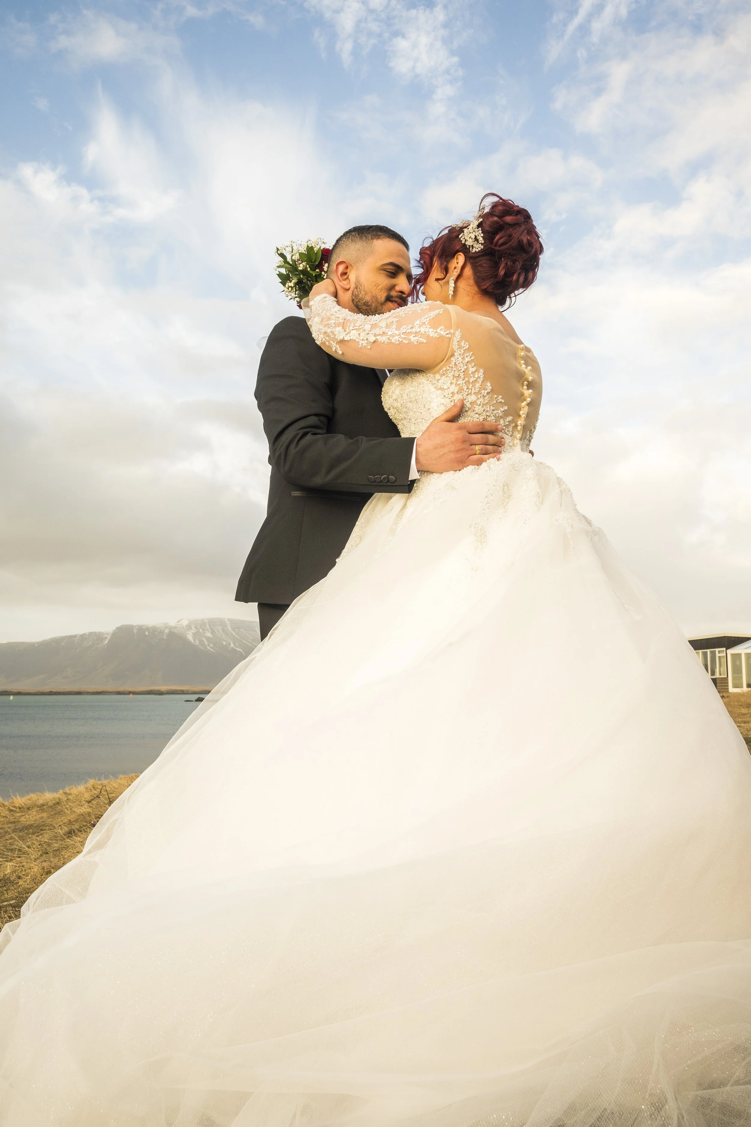 A bride and groom embrace with lakes, mountains, and a cloudy sky in the background on their wedding day.