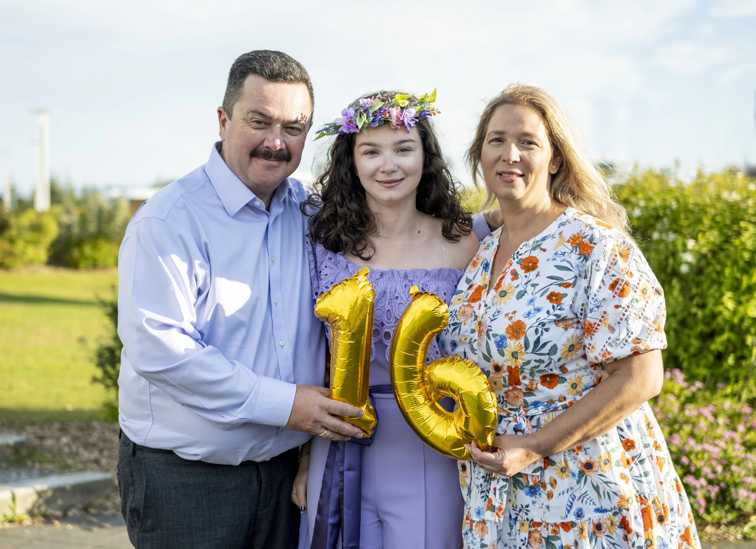 Family celebrating a 16th birthday outdoors, holding gold balloons shaped as the numbers 1 and 6, with the teenage girl wearing a flower crown and purple outfit, surrounded by greenery and a bright sky.