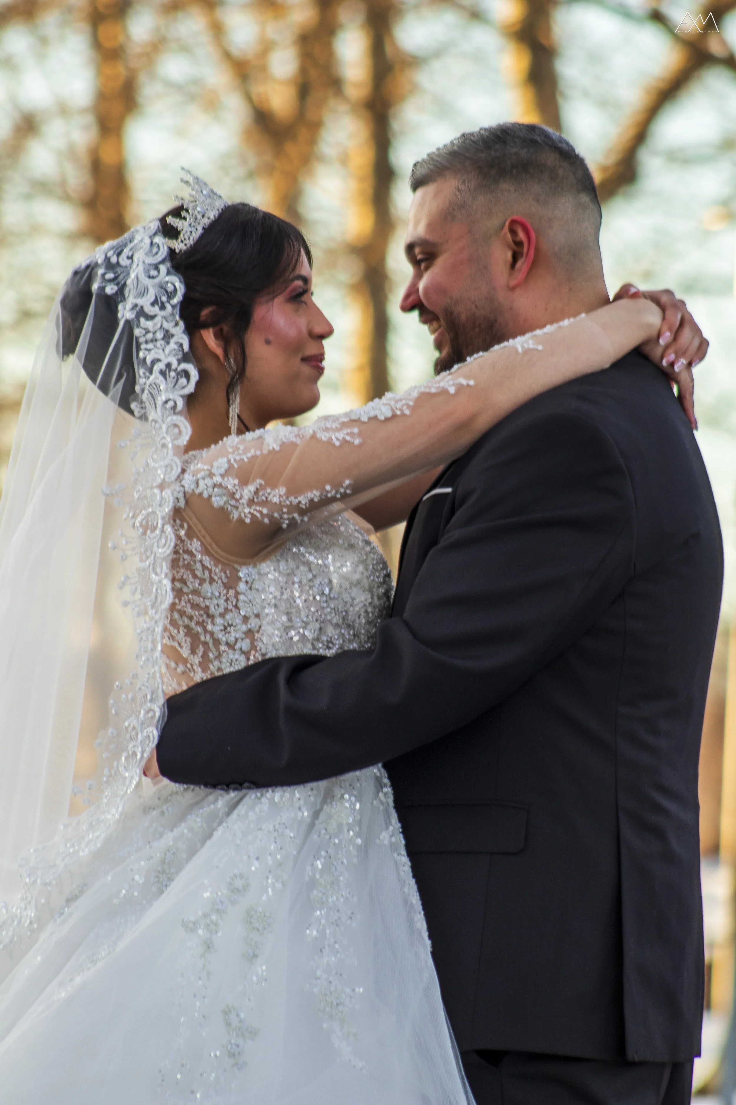 A bride and groom are embracing and looking into each other's eyes during their wedding ceremony outdoors, with trees in the background.
