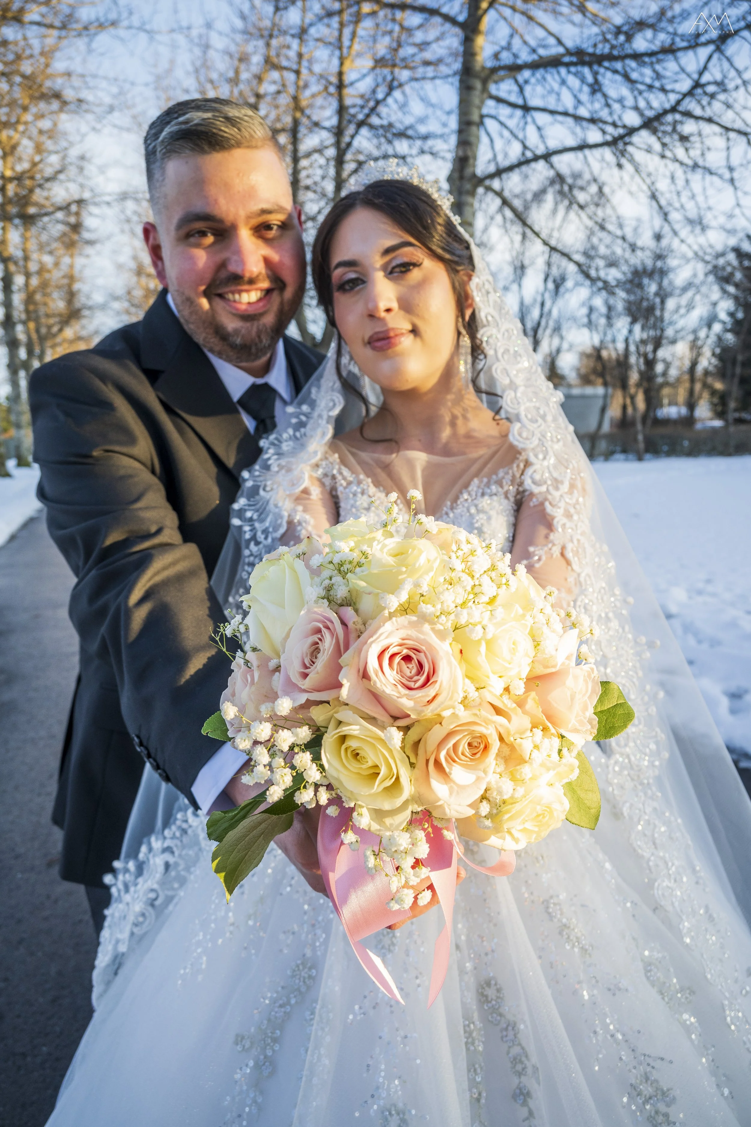 A bride and groom smiling and posing outdoors in a winter setting, with leafless trees and snow on the ground, the bride wearing a white lace wedding dress and the groom in a black suit holding a bouquet of light pink, white, and cream roses with gre