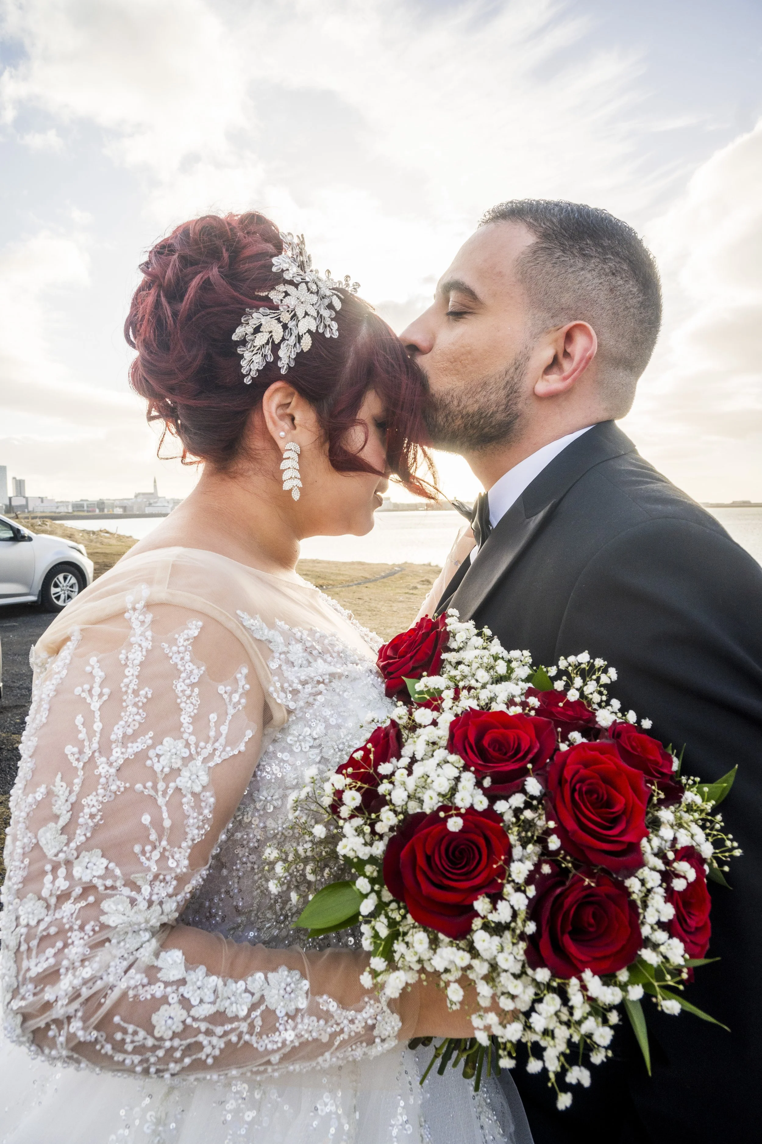 A bride and groom sharing a kiss outdoors, with a bride in a white lace wedding dress with floral details and a groom in a black tuxedo, holding a bouquet of red roses and white flowers, against a cloudy sky and waterfront background.