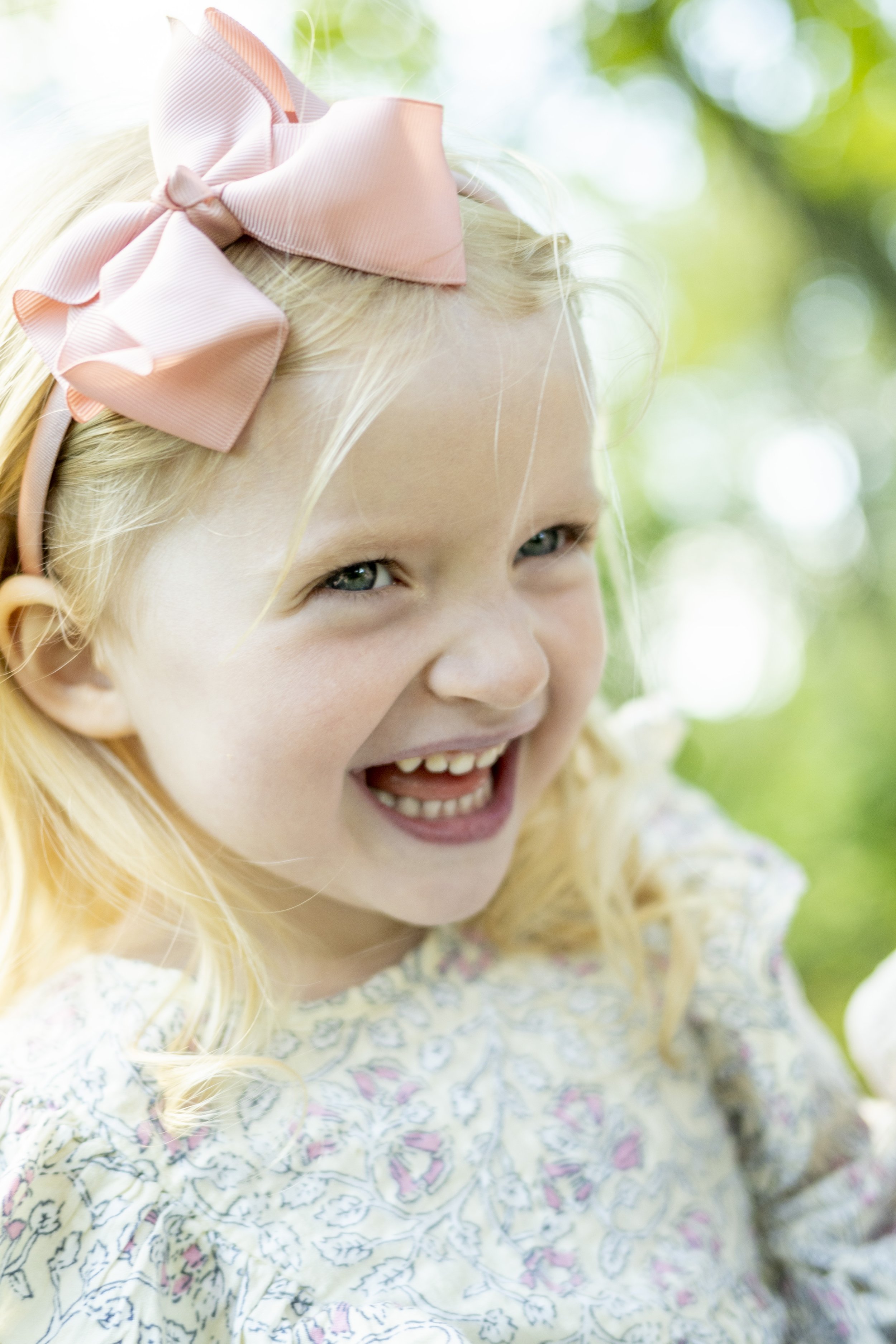 A young girl with blonde hair, smiling and showing her teeth, wearing a pink hair bow and a patterned dress outdoors with green foliage in the background.
