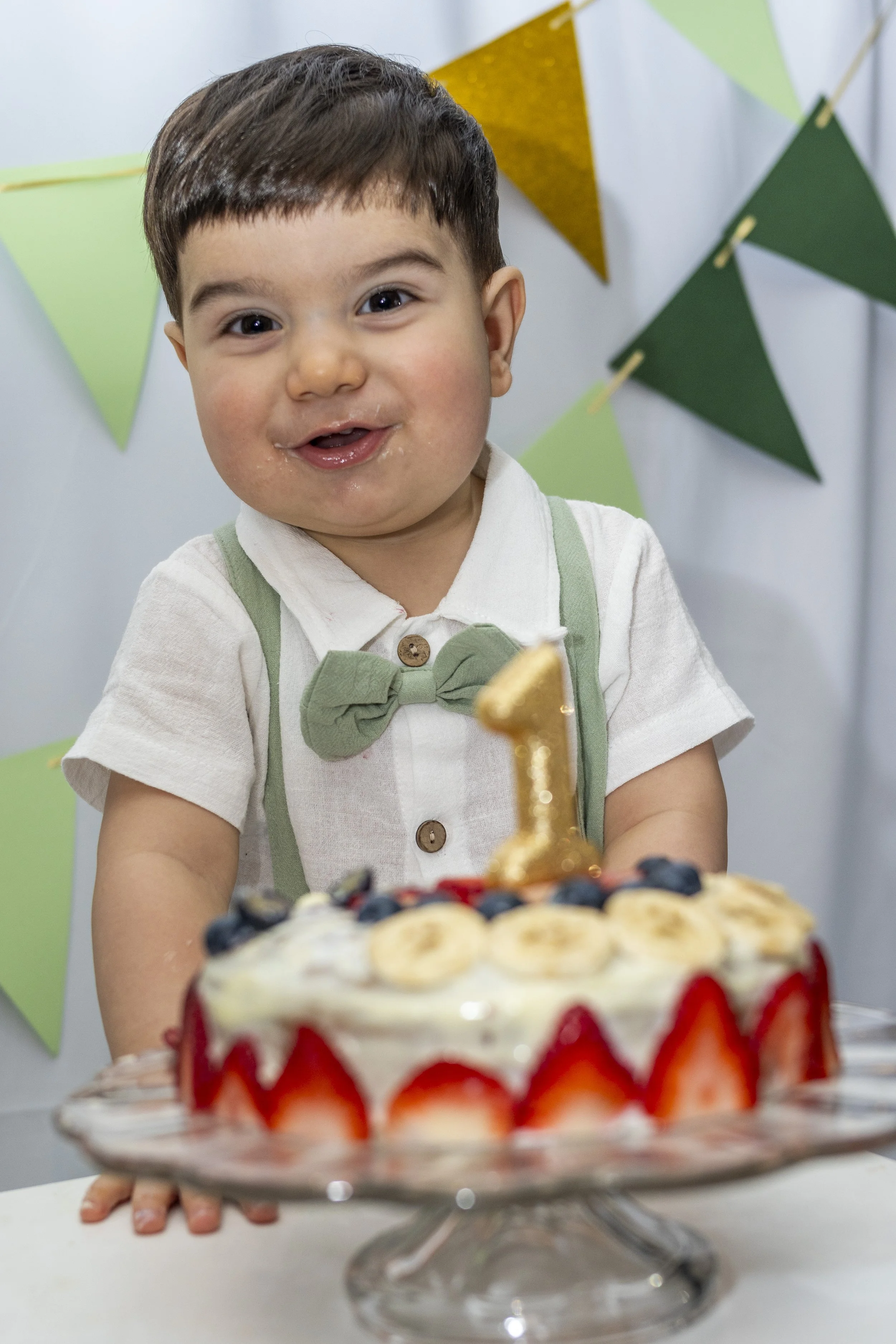 A young boy with short dark hair and light skin, wearing a white shirt and green suspenders with a green bow tie, celebrates his birthday with a cake decorated with strawberries, bananas, blueberries, and a gold number 1 candle. The background featur