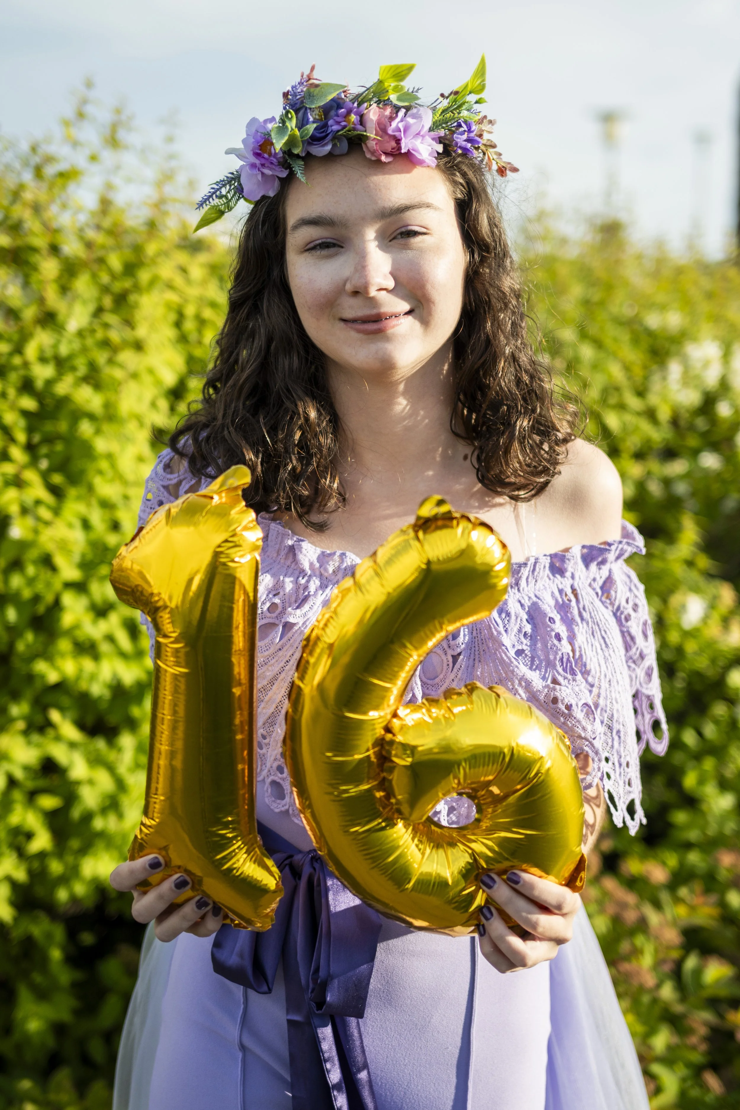 Young woman celebrating her 16th birthday outdoors, wearing a flower crown, holding gold balloons forming the number 16, and smiling.