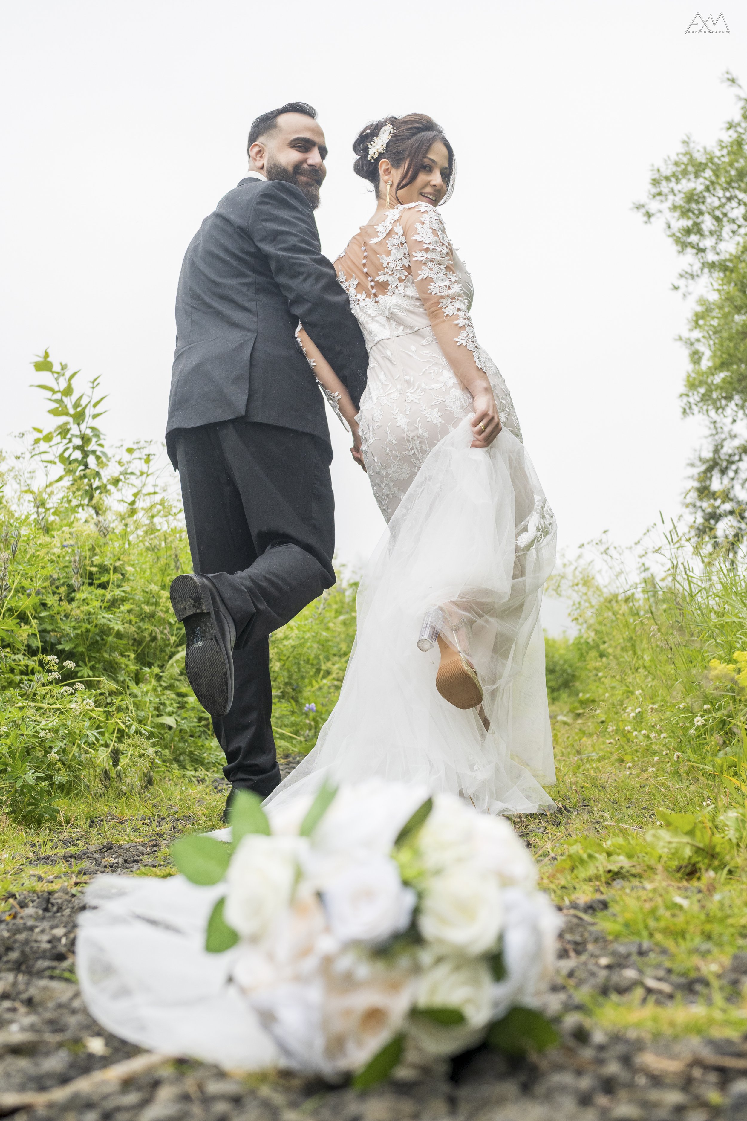 A smiling bride and groom holding hands outdoors, with a bouquet of white flowers with green leaves in front of them. The bride is wearing a lace wedding dress with floral embroidery and a floral hair accessory. The groom is wearing a black suit and 