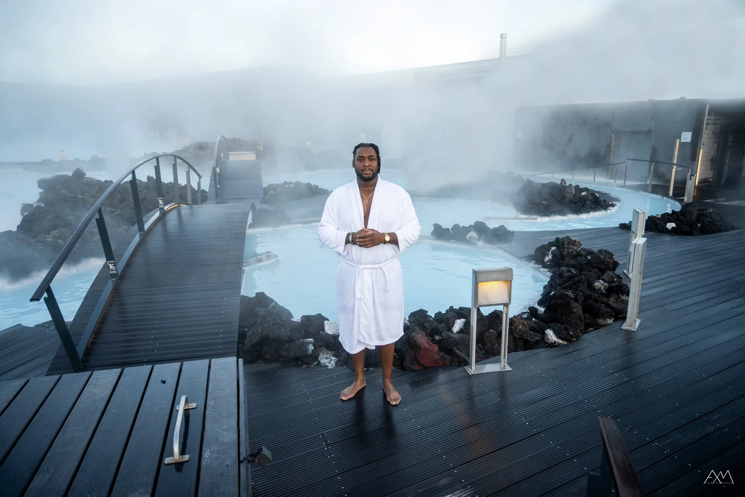 Man in a white bathrobe standing barefoot on a wooden deck by a hot spring, with steam rising and rocks around the water, in an outdoor spa setting.