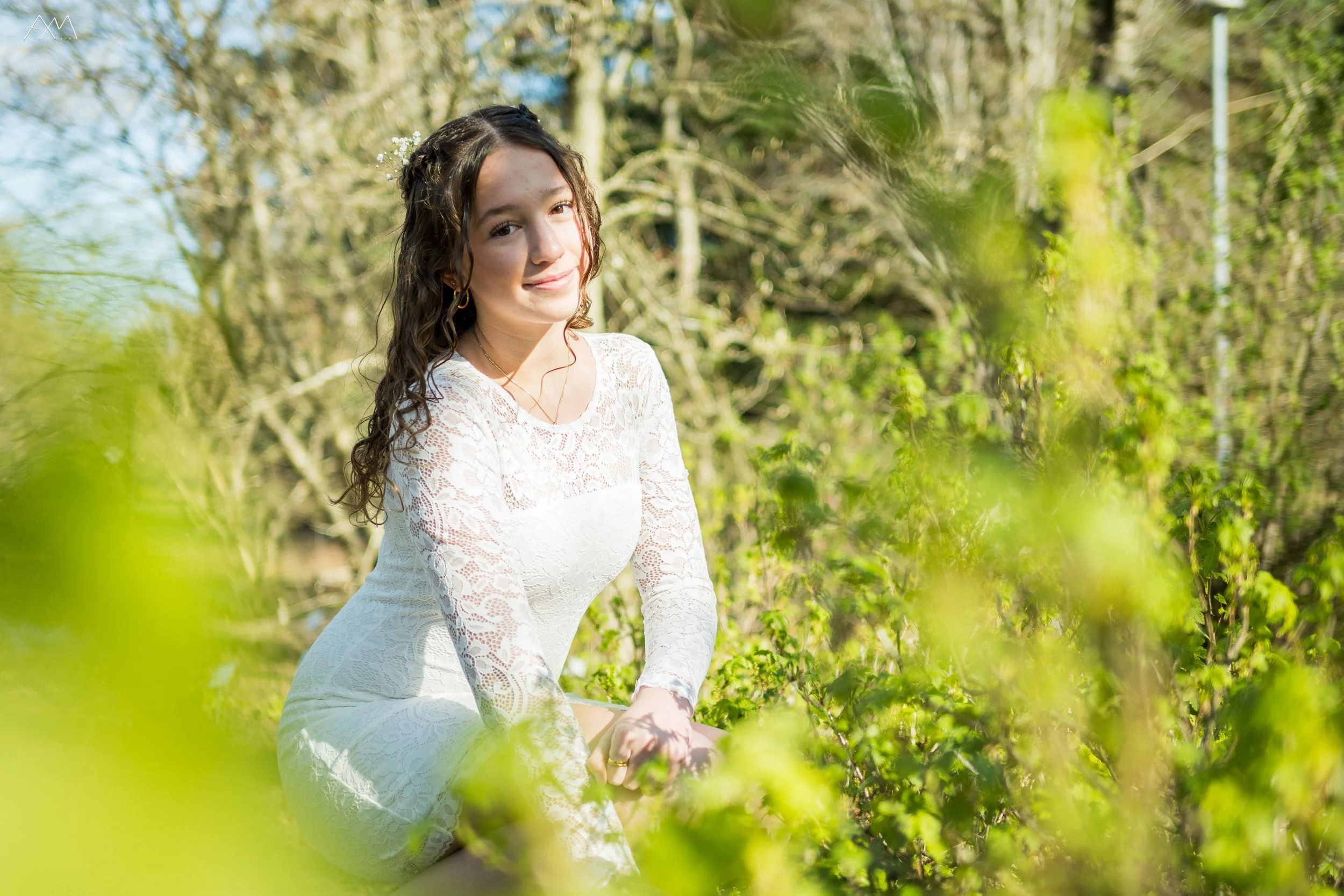 A young woman with curly brown hair, wearing a white lace dress and gold earrings, sitting outdoors among green foliage and trees, smiling softly at the camera on a sunny day.