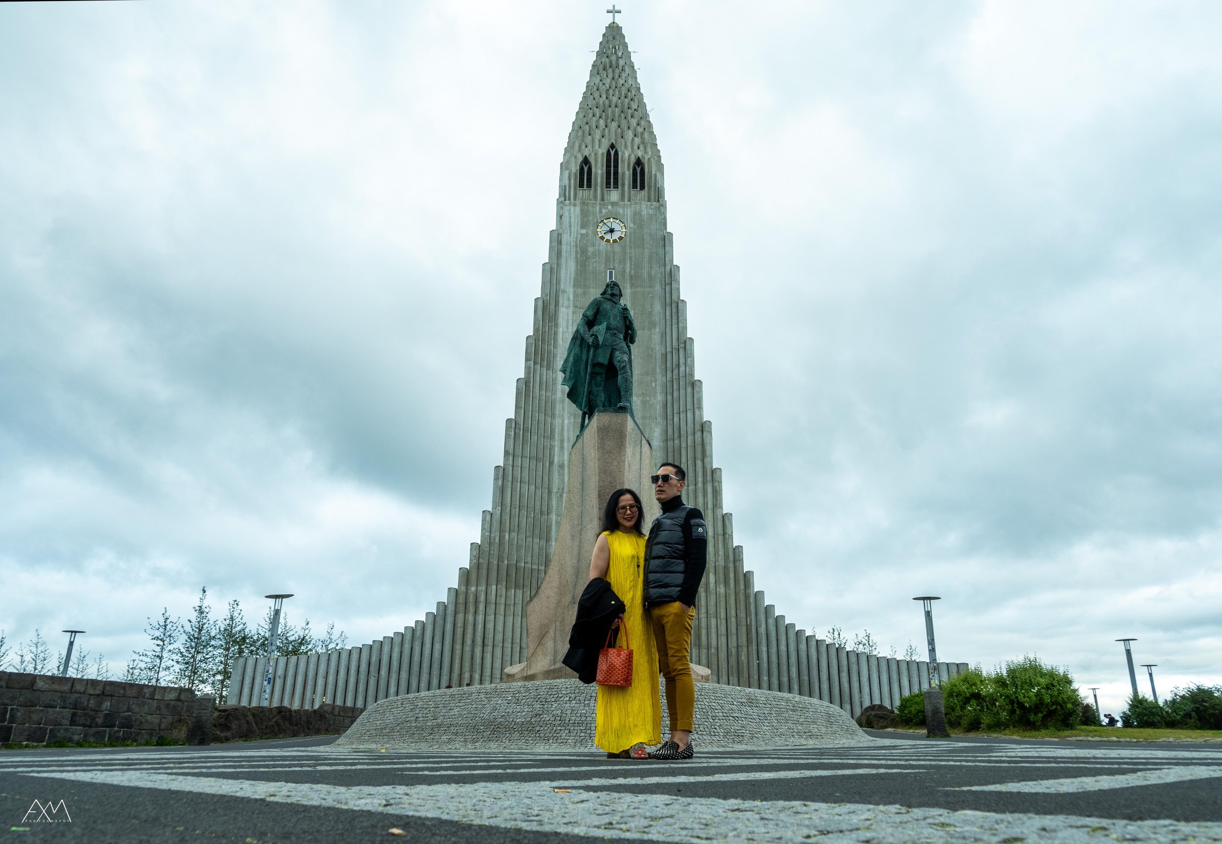 Two people standing in front of a large, gothic-style church Hallgtimskirkja in Iceland with a tall, pointed spire and a statue of a man below the spire. The sky is overcast.
