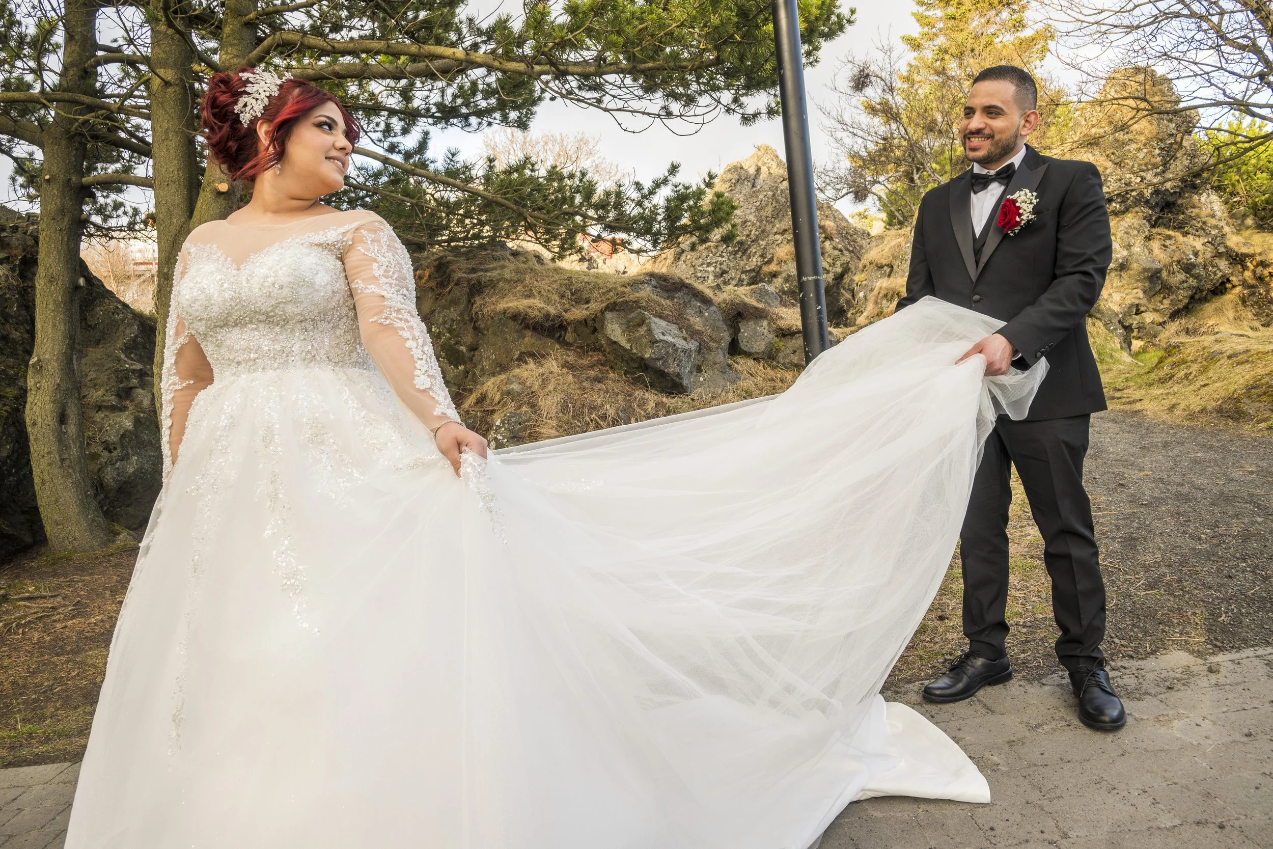 A bride and groom outside during the day, with the groom holding the bride's wedding dress train. The bride is wearing a white lace wedding gown with long sleeves and a floral hairpiece. The groom is dressed in a black tuxedo with a white shirt and b
