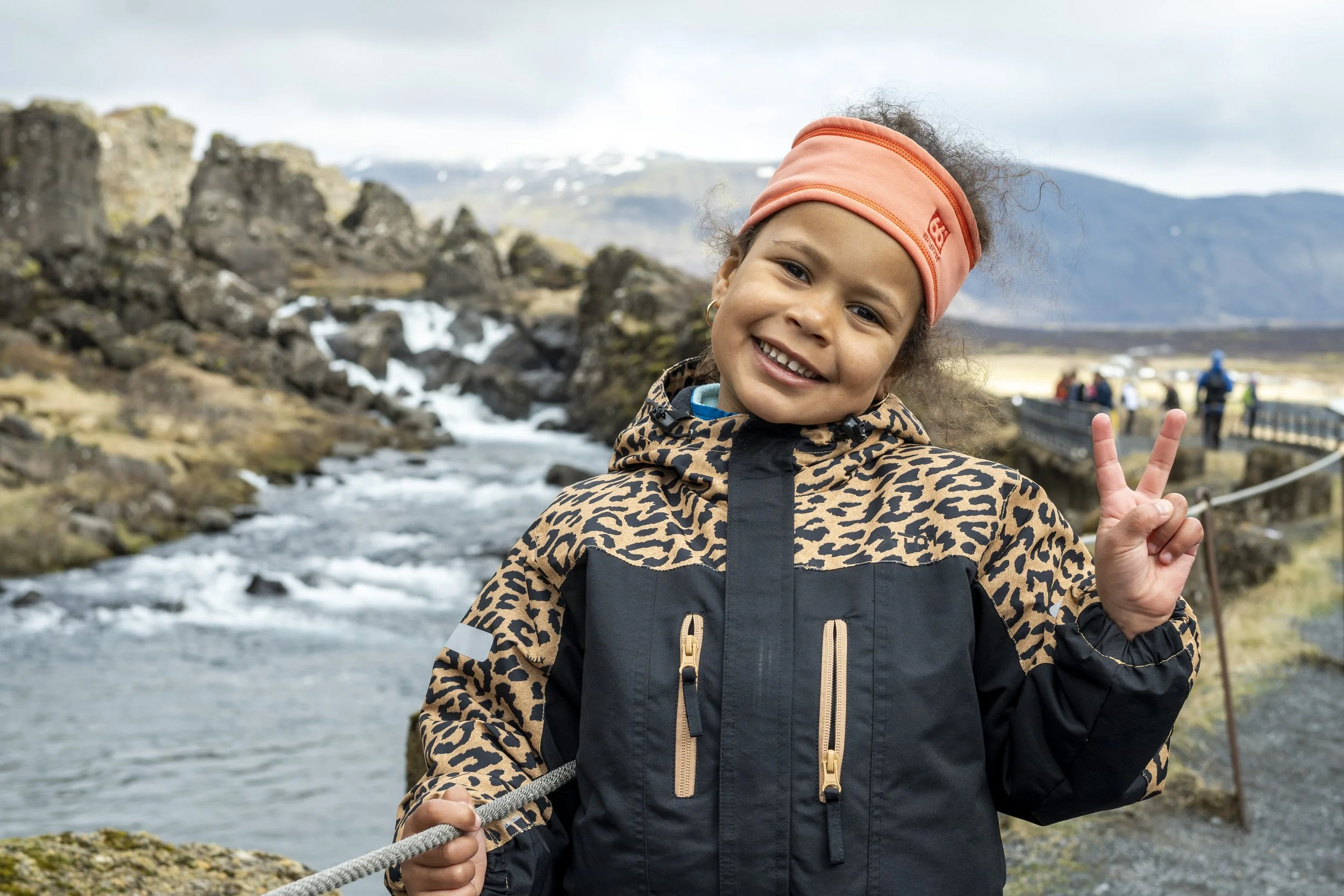A young girl smiling and making a peace sign, wearing a leopard-print jacket and a peach-colored headband, standing outdoors near a river with a waterfall and mountains in the background.