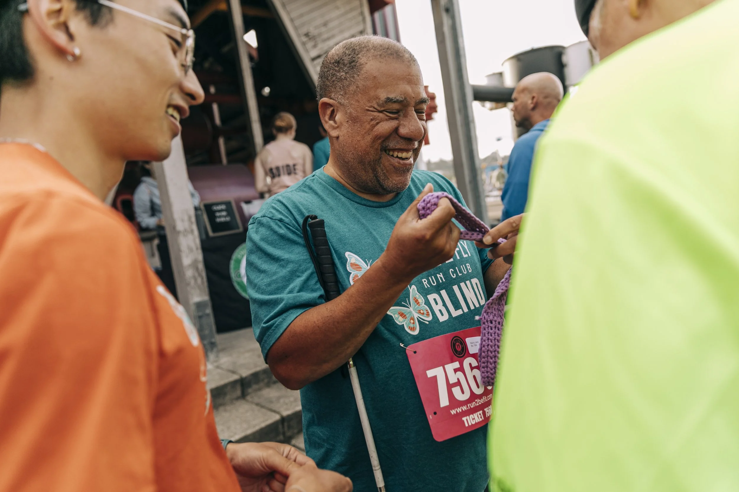 People participating in a marathon or race, with a focus on a smiling man in a blue shirt holding a race bib, surrounded by others in colorful attire.