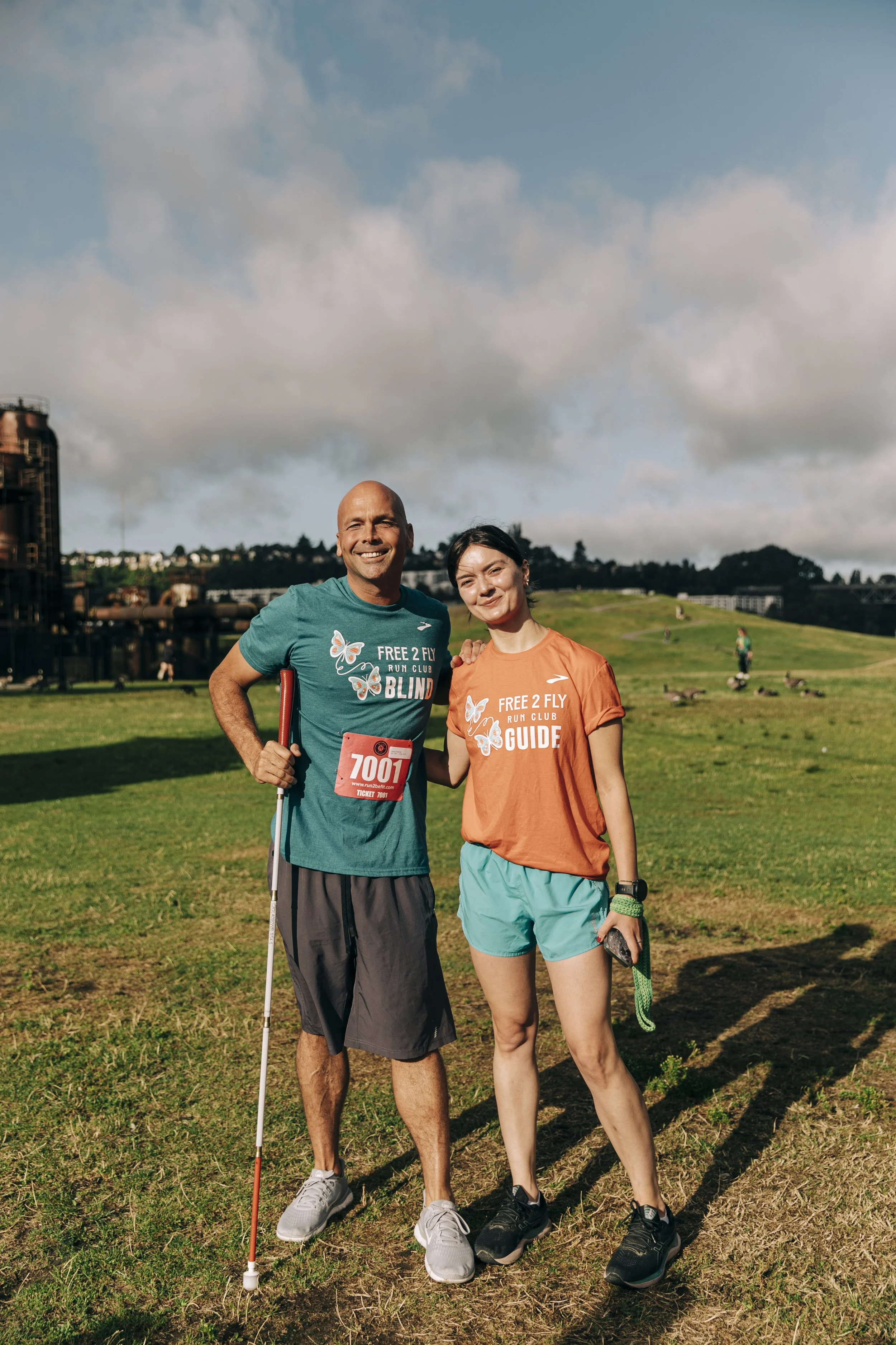 Two smiling runners, a man and a woman, posing in a park with a grassy hill and cloudy sky in the background. Both are wearing athletic attire with matching event T-shirts, with bib numbers and accessories.