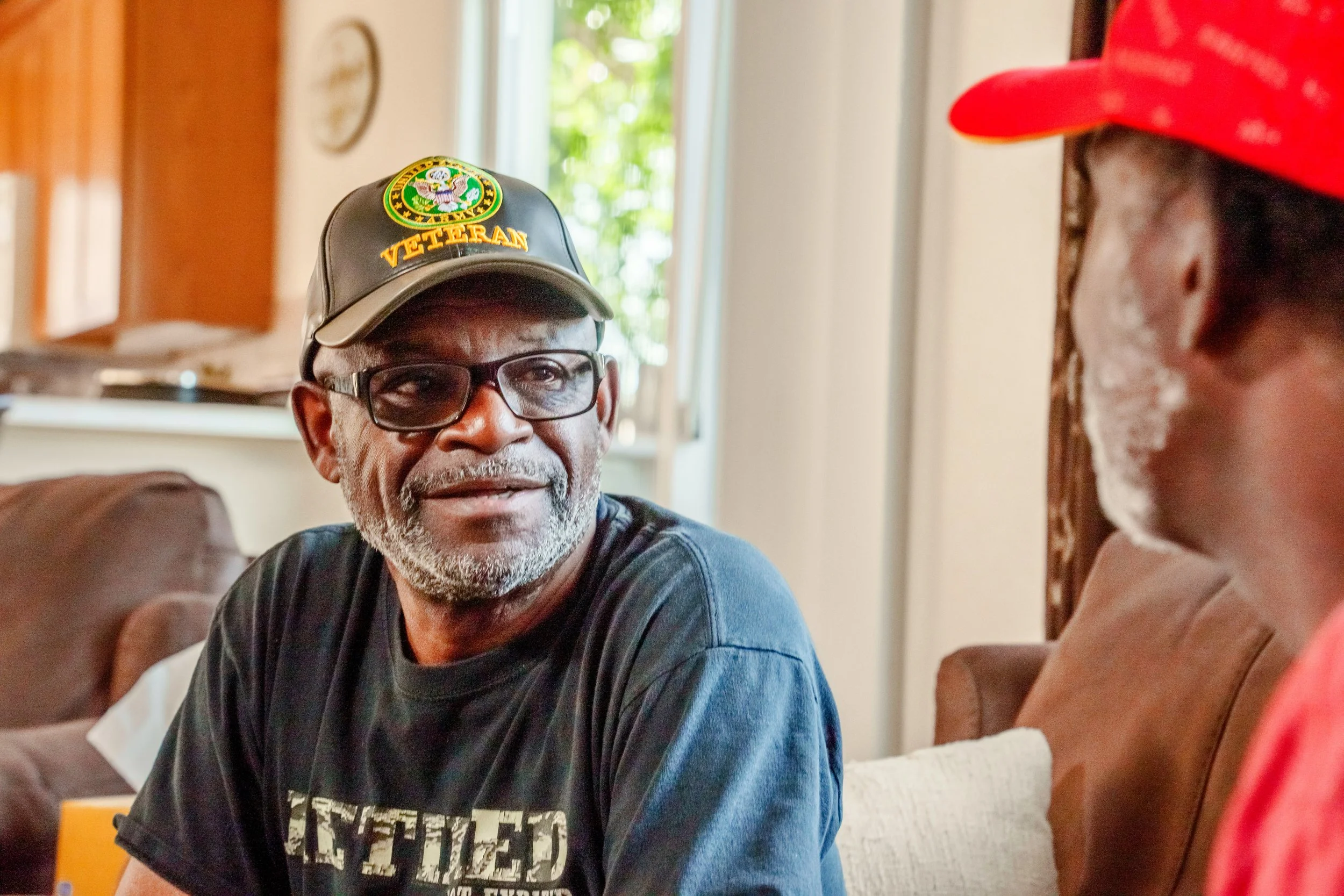 An elderly man with glasses, a gray beard, wearing a black t-shirt and a veterans hat, sitting on a couch and smiling at another person wearing a red cap.