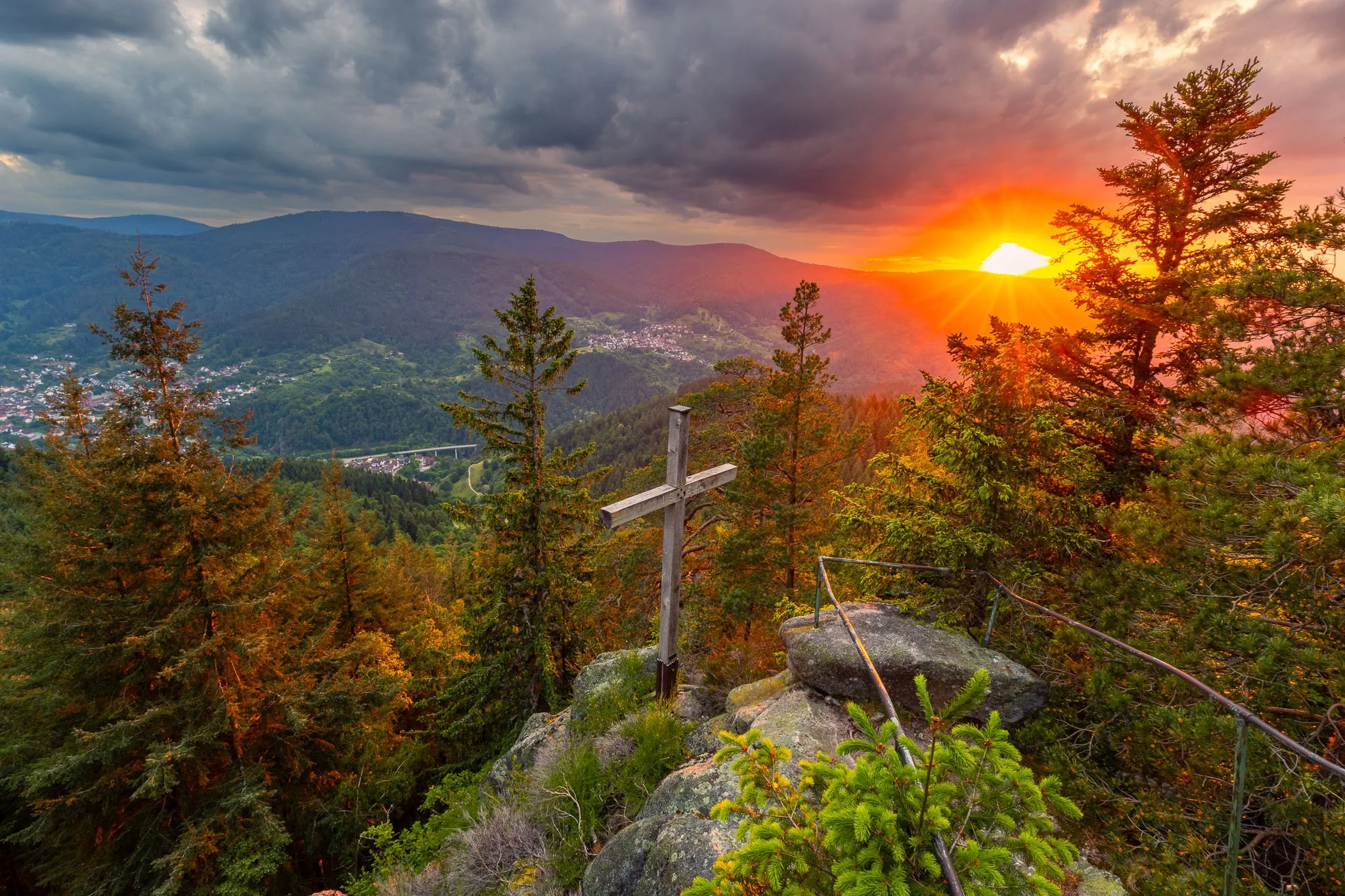 Bergpanorama bei Sonnenuntergang mit einem Holzkreuz auf einem Felsen im Vordergrund, umgeben von Bäumen, mit einer Stadt im Tal und Bergketten im Hintergrund.