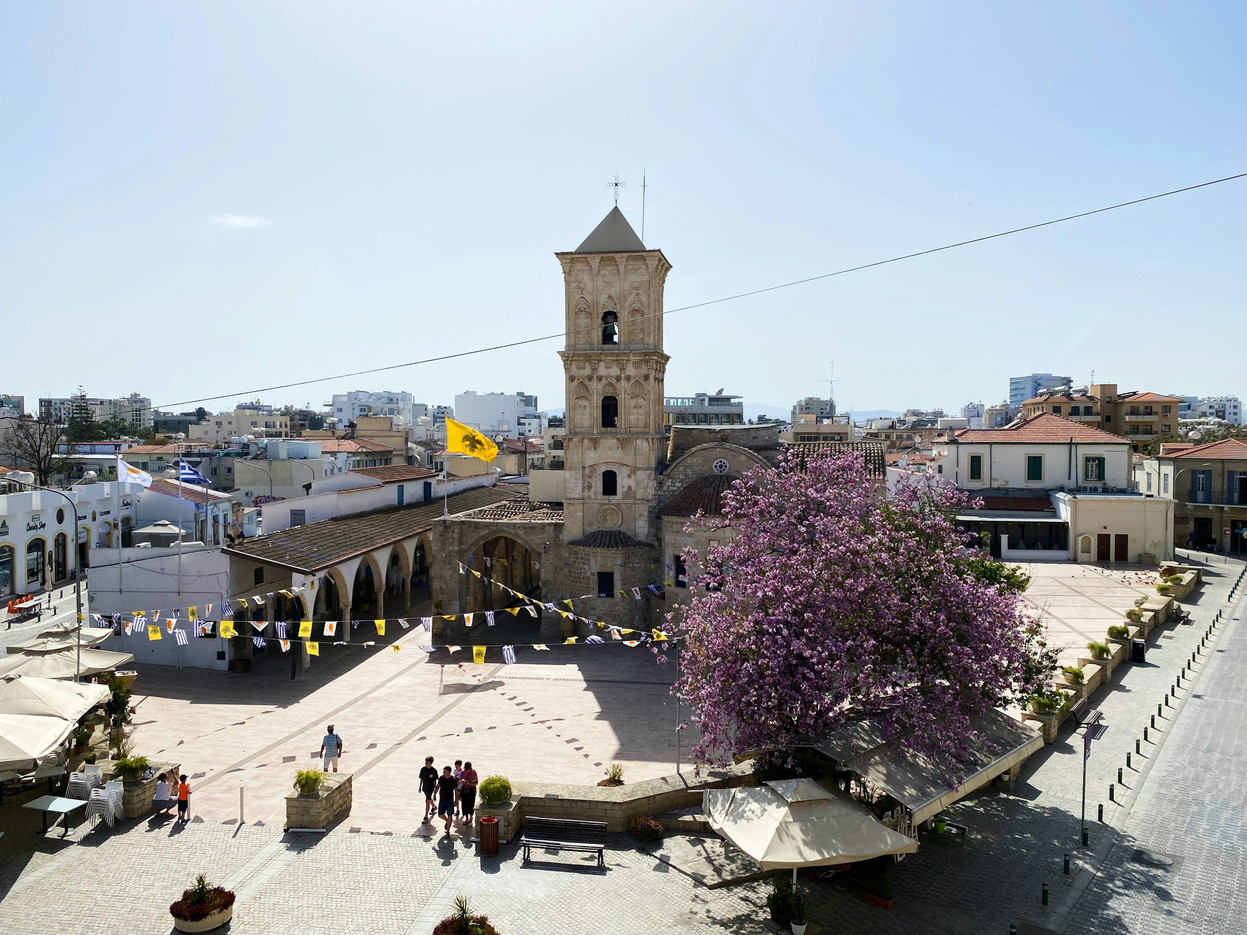 A historic church with a bell tower in a town square, decorated with yellow and Greek flags, with blooming pink trees and outdoor seating areas.