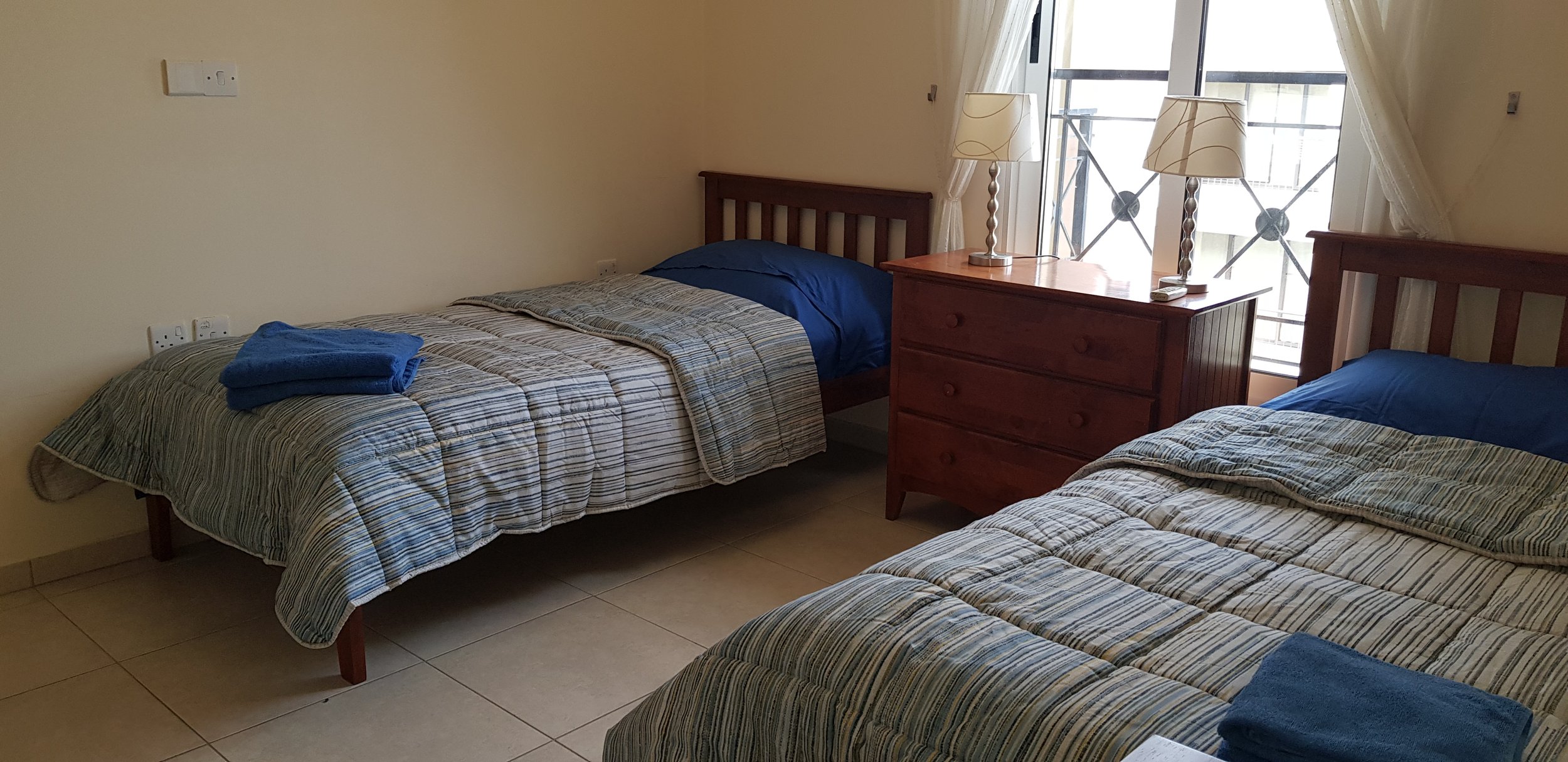 A bedroom with two twin beds with striped bedspreads and blue pillows, a wooden nightstand with two lamps, and a window with curtains and a balcony railing outside.