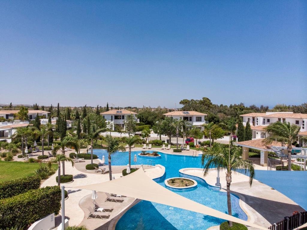 Resort swimming pool area with lounge chairs, palm trees, and surrounding modern white buildings under a clear blue sky.