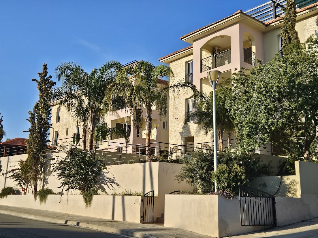 Multi-story residential building with balconies, surrounded by trees and palm trees, concrete wall, against a clear blue sky.