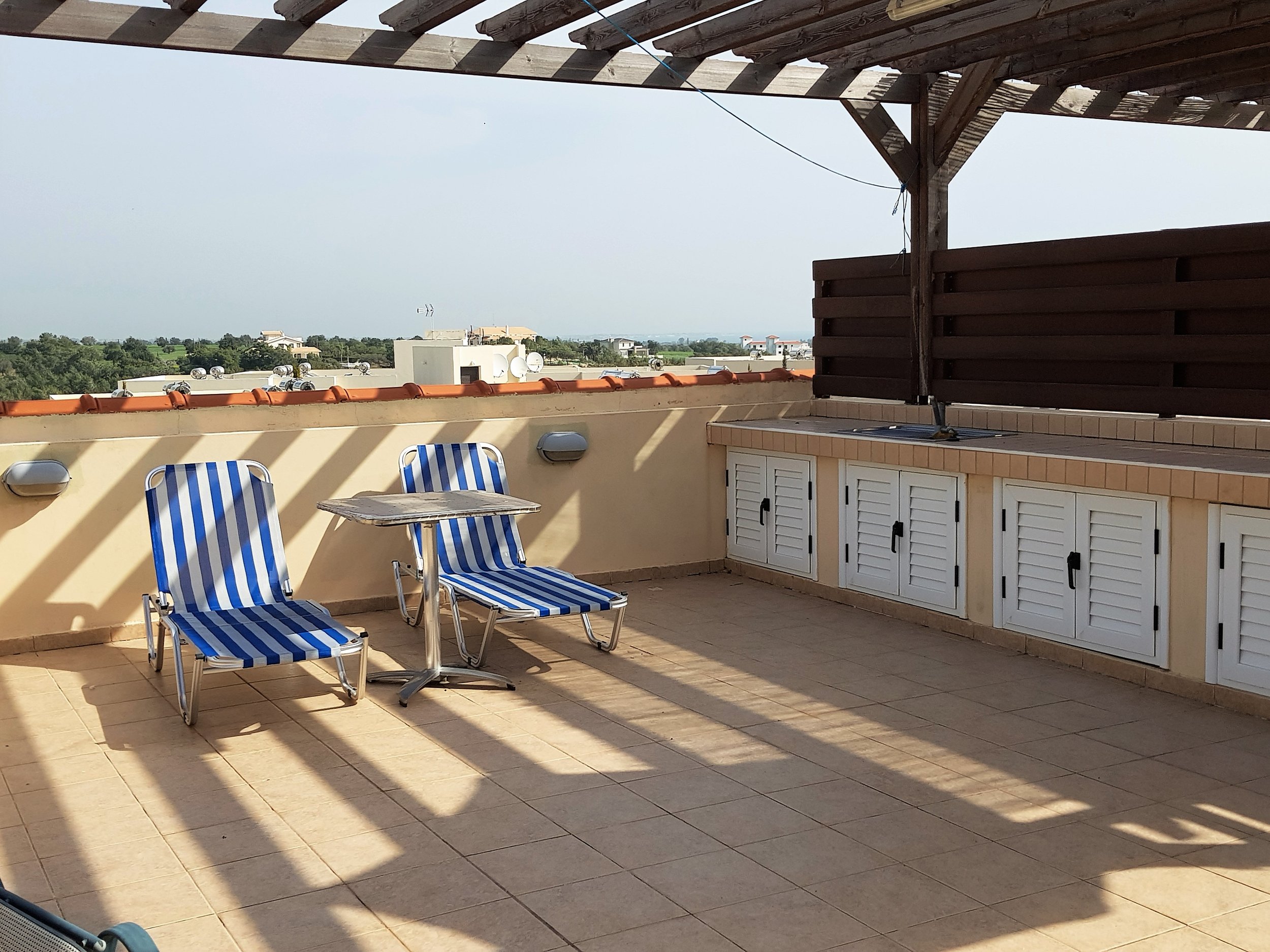 Rooftop terrace with two blue and white striped lounge chairs, a small round table, wooden pergola, and white storage cabinets in the corner. There's a view of rooftops and greenery in the distance.