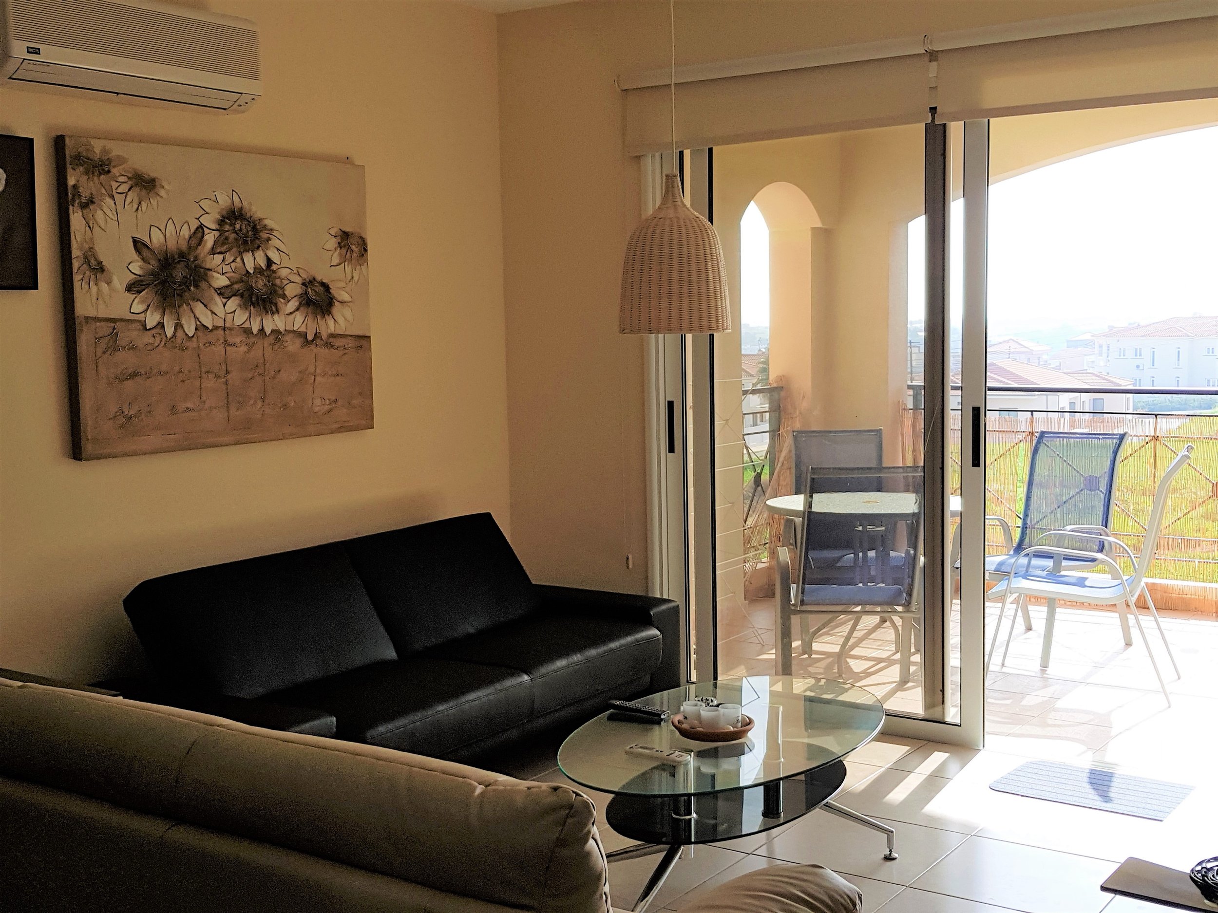 Living room with black leather sofa, beige armchair, glass coffee table with tea setup, artwork with sunflowers on the wall, view of balcony with outdoor chairs and table, sliding glass door leading outside, and bright natural light.