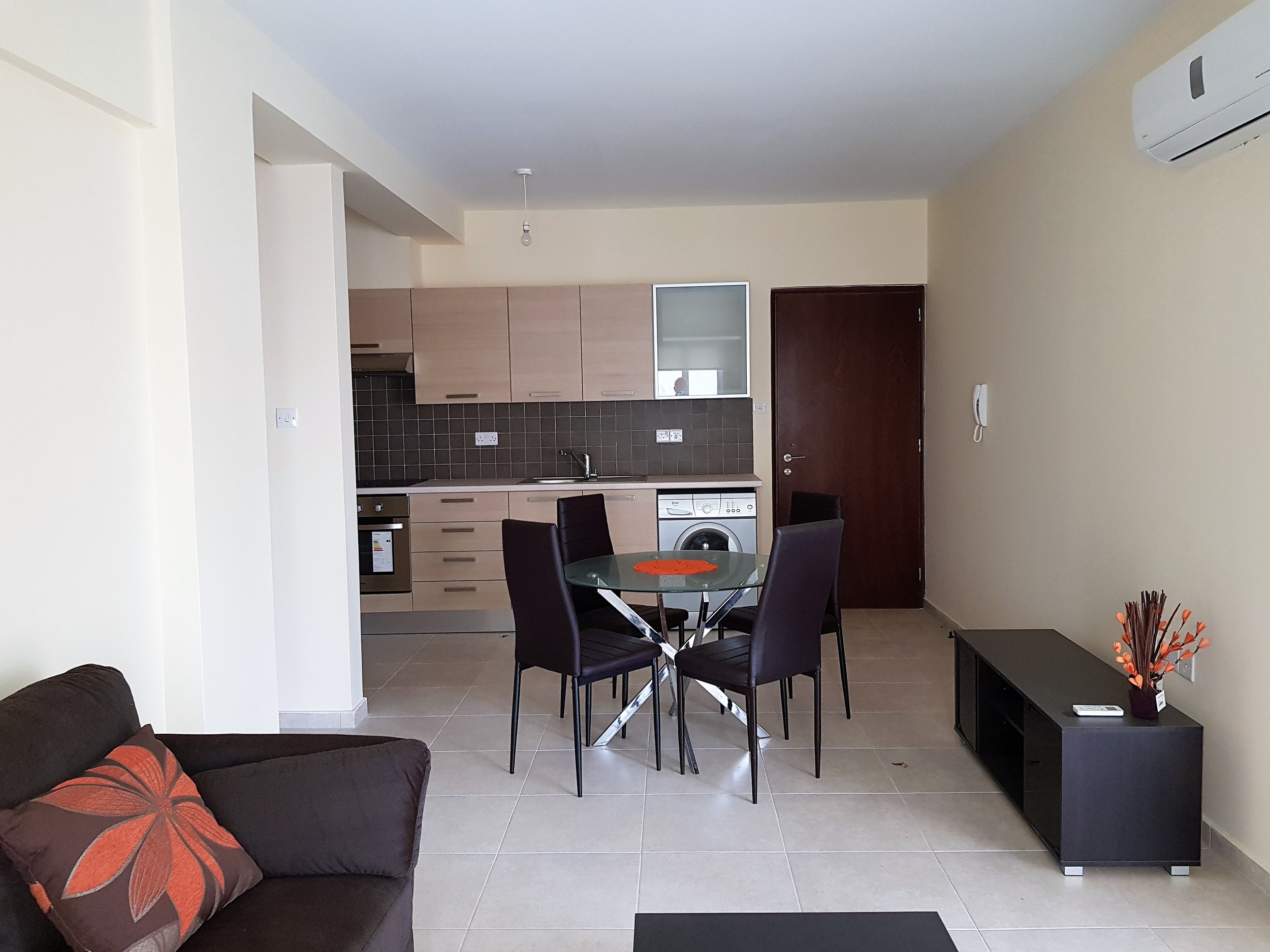 Open-concept living space with a sofa, dining table with four chairs, kitchen with beige cabinets, gray backsplash, washing machine, and wall-mounted air conditioner.