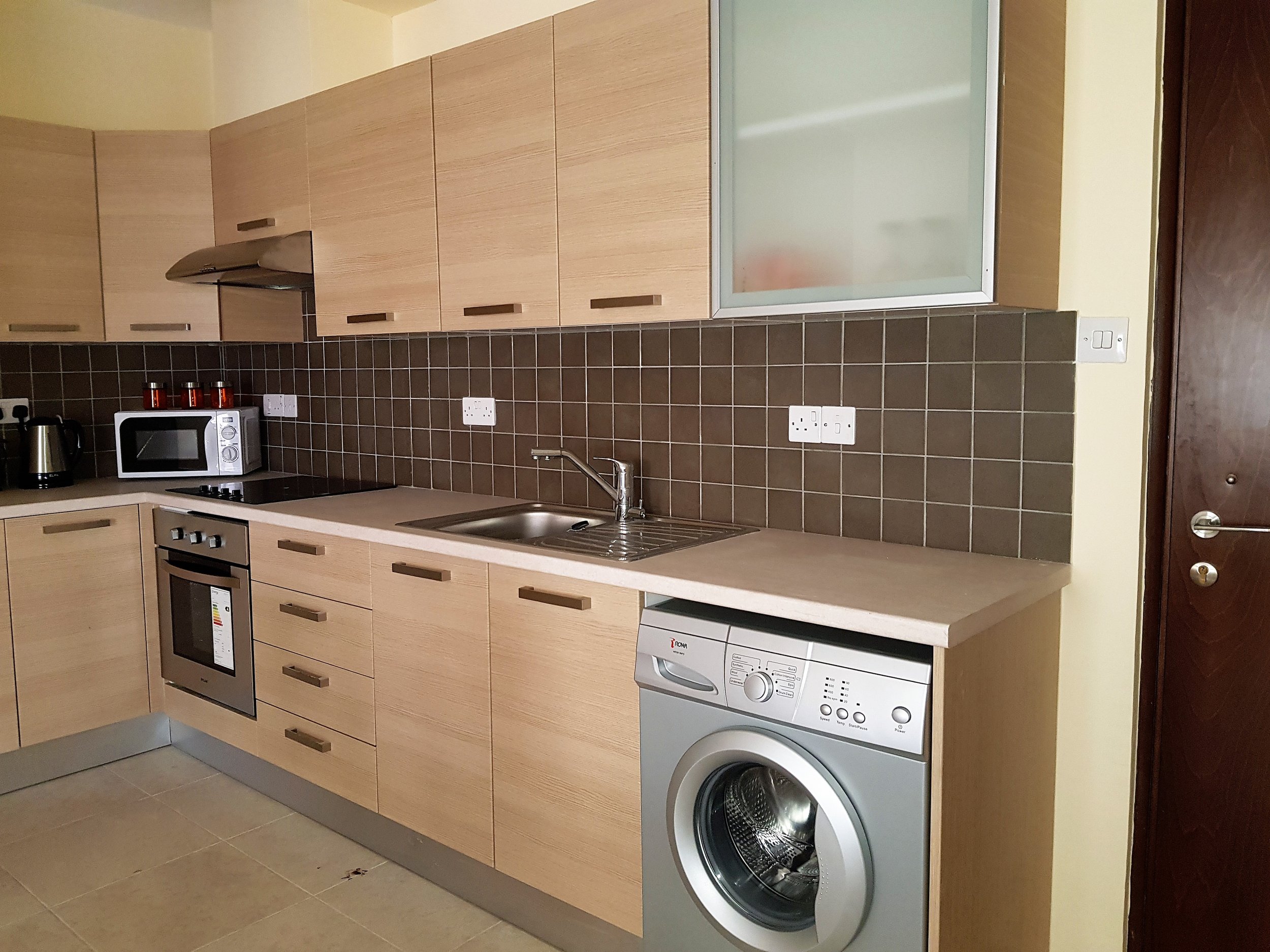 Modern kitchen with wooden cabinets, brown tiled backsplash, microwave, kettle, oven, sink, and washing machine.