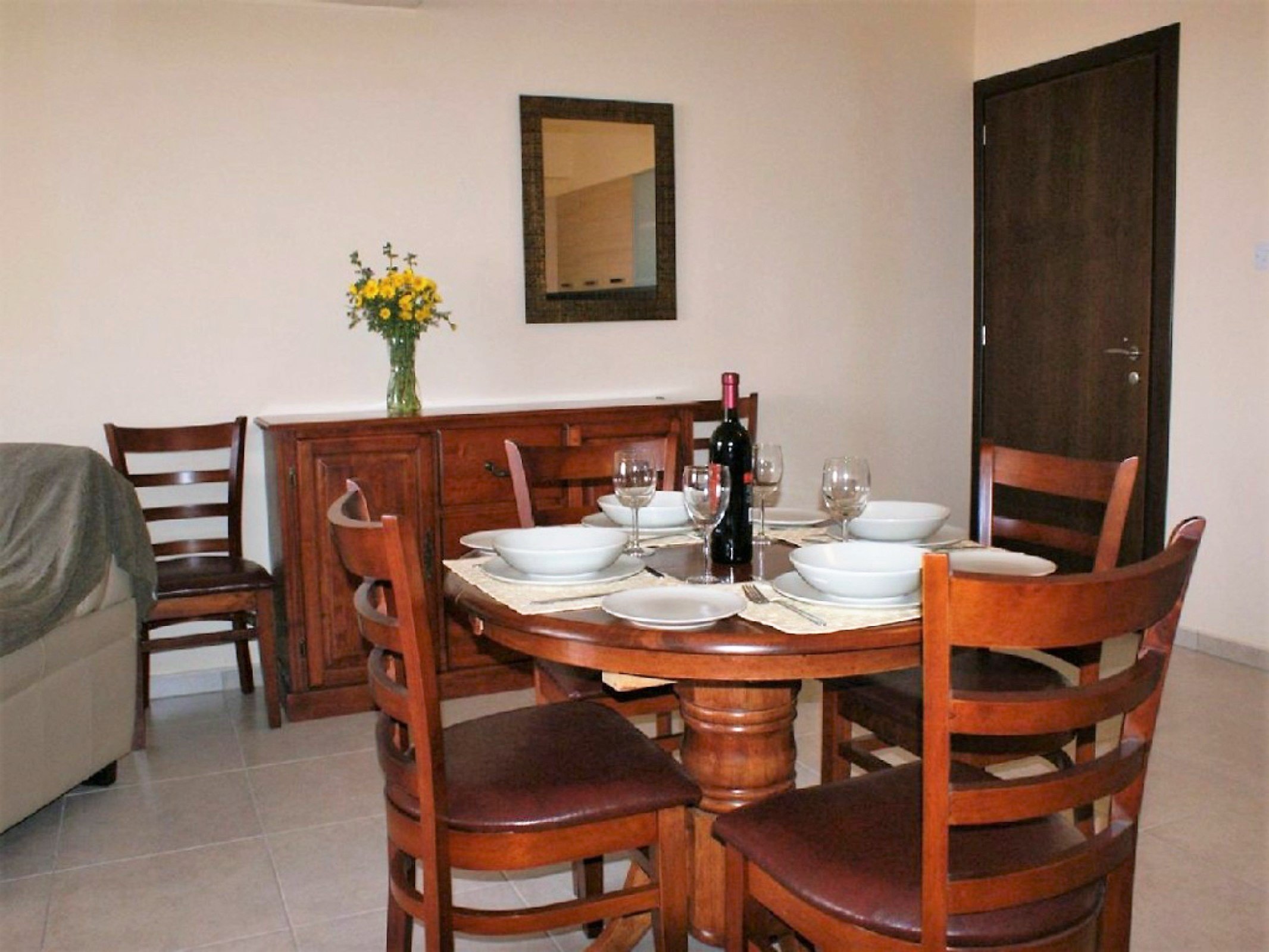 Dining table set with white bowls, wine glasses, a bottle of wine, and cutlery, in a room with a wooden sideboard, a mirror, and a vase of yellow flowers.