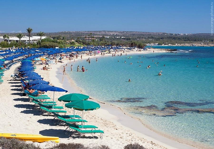 Beach scene with turquoise water, white sand, colorful umbrellas, and people swimming and sunbathing.
