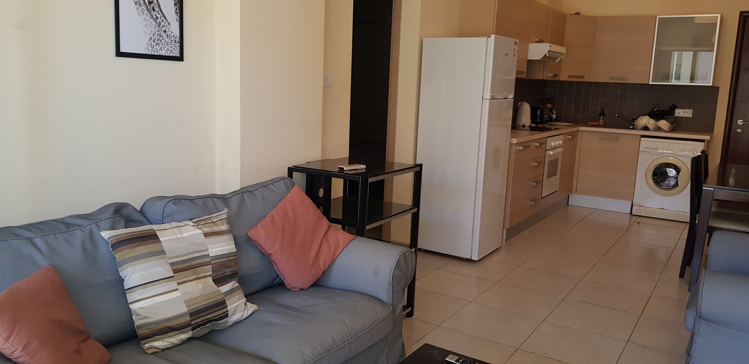 Living room with a blue sofa, three cushions, a small black side table, and an open kitchen with a white refrigerator, washing machine, stove, and various kitchen appliances.