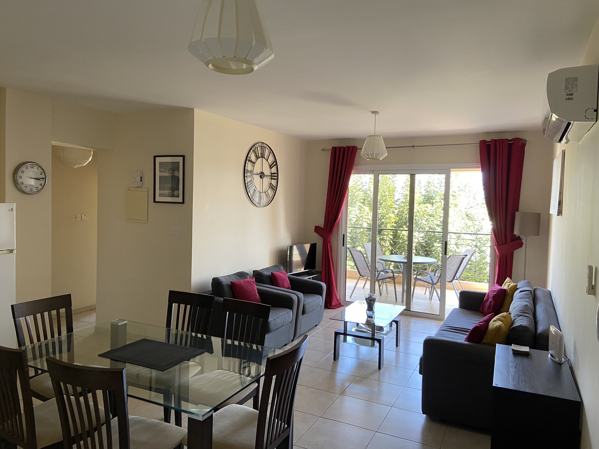 Living room with two gray sofas with red and yellow pillows, glass coffee table, dining table with black chairs, balcony with table and chairs, large sliding door, curtains, wall clocks, wall art, TV, air conditioning, and a ceiling light.
