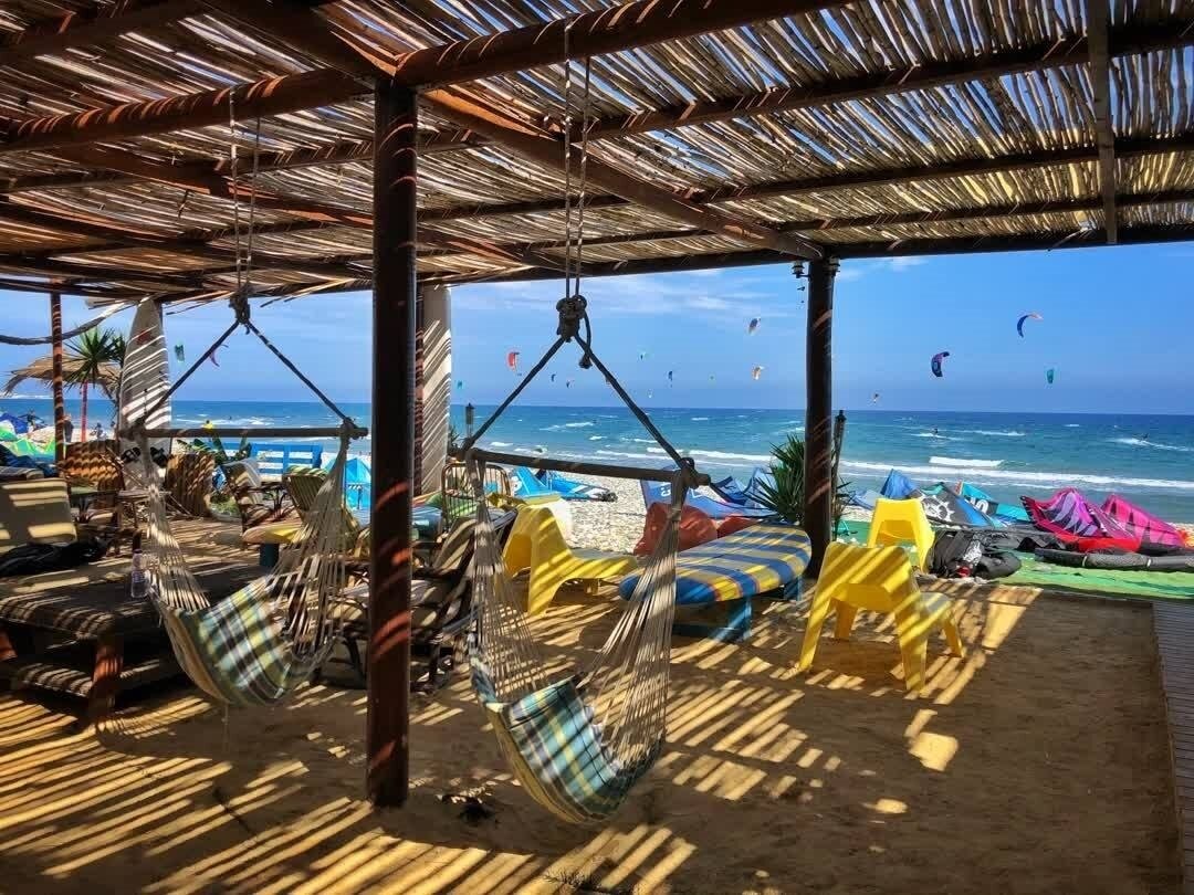 Beachside cabana with hammocks, colorful lounge chairs, and kitesurfers in the ocean under a sunny sky.