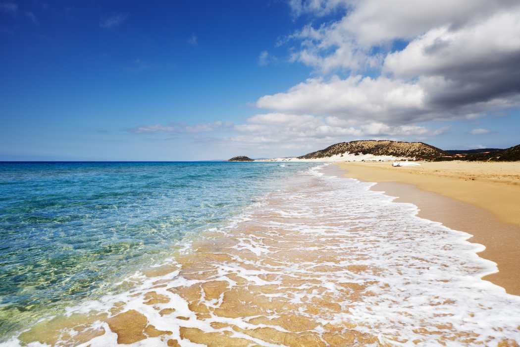 Scenic beach with golden sand, clear blue ocean, partly cloudy sky, and distant hills.
