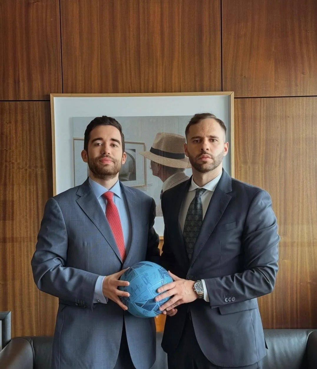 Dois homens de terno segurando uma bola azul assinada, posando para a câmera em um ambiente interno, com uma pintura de fundo.