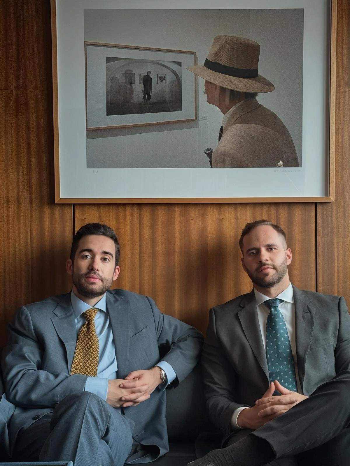Dois homens de terno sentados em um assento, com uma moldura de quadro na parede de madeira ao fundo. No quadro, uma fotografia em tons de cinza de uma pessoa observando obras de arte em uma galeria.