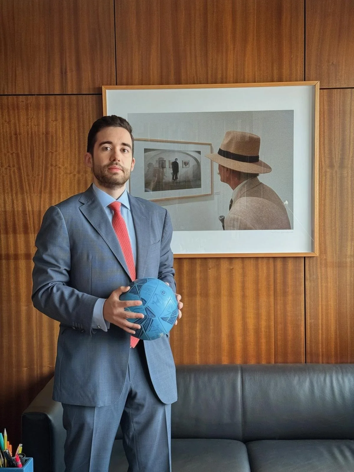 Homem bem vestido com terno cinza, camisa azul e gravata vermelha segurando uma bola azul de futebol, em um escritório com parede de madeira e quadro na parede.