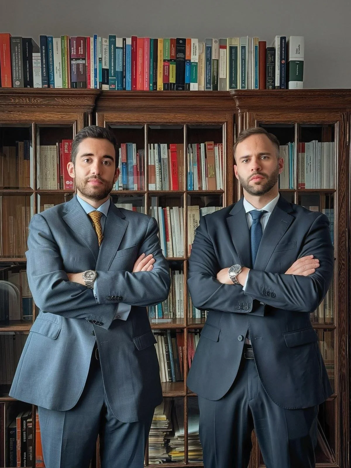 Dois homens de terno com braços cruzados, um na esquerda e outro na direita, posando na frente de uma estante de livros de madeira.