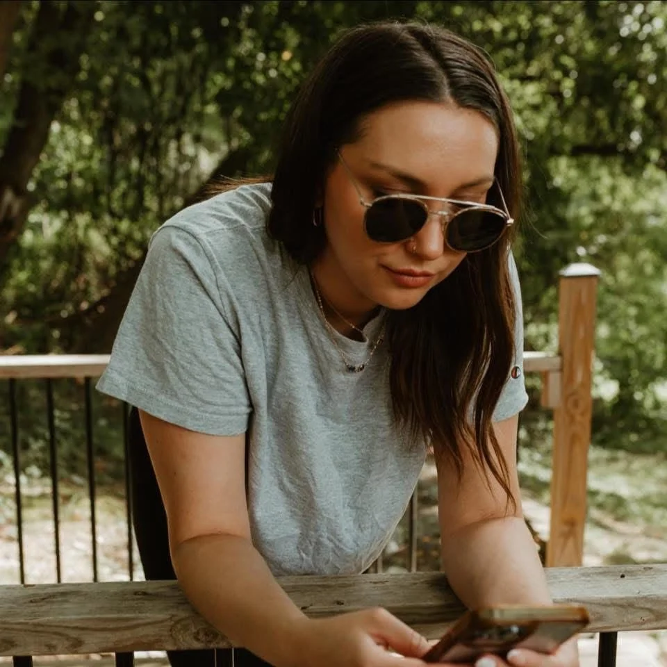 Young woman with dark hair and sunglasses wearing a gray t-shirt, leaning over a wooden table and looking at her phone outdoors surrounded by green trees and sunlight.