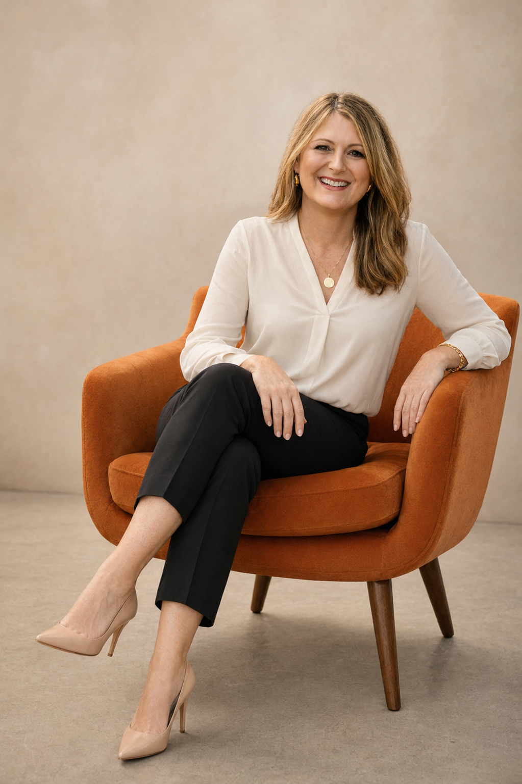 A woman with shoulder-length light brown hair sitting on an orange armchair, smiling, wearing a white blouse, black pants, beige high heels, and gold jewelry, against a plain beige background.